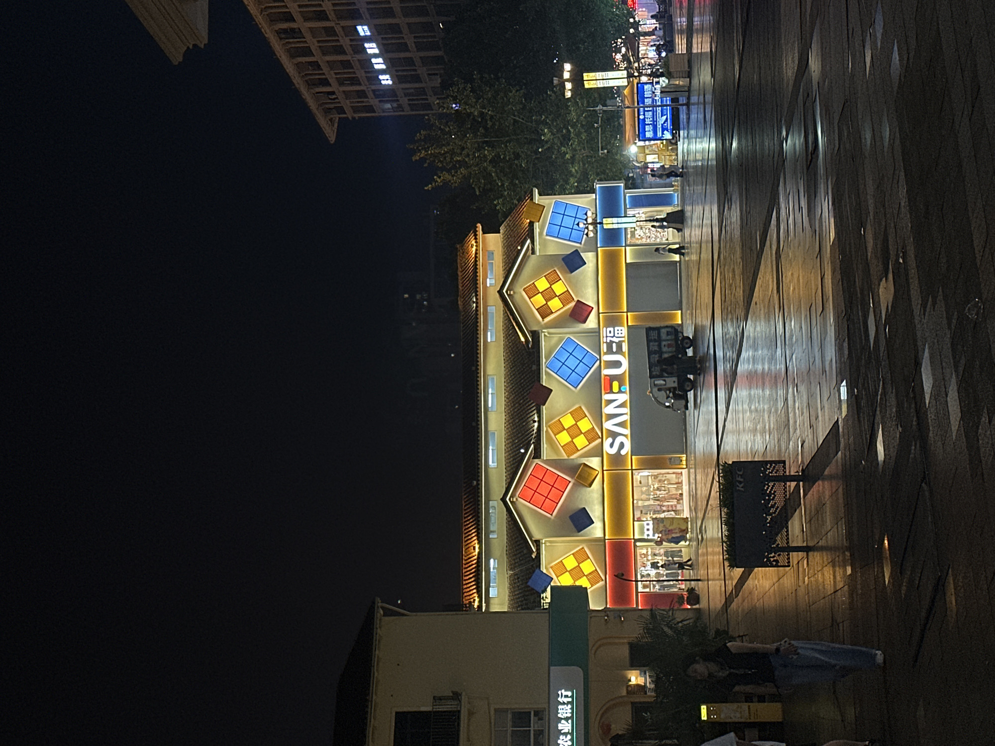 In the city of Chongqing High-tech Zone, China, at night, a wide, wet plaza is illuminated by streetlights and the neon signs of businesses. The most prominent structure is a building with a pitched roof, decorated with large, colorful, square panels, some featuring a checkered pattern and others solid blue. The building's facade displays the illuminated signage "SAN-U" in large, white, block letters. To the left, another building with a green awning displays Chinese characters. In the foreground, a woman in a dark top and light-colored pants walks across the wet pavement, her reflection visible in the puddles. Several other individuals are scattered across the plaza, some appearing to be shoppers entering or exiting stores. A small, dark vehicle with a white upper section is parked in front of the "SAN-U" building. The plaza floor is paved with large, dark tiles, some of which are slick with water, creating reflections of the surrounding lights. In the background, a tall, modern building with many windows is visible. The overall scene depicts a lively commercial area at night, with the wet surfaces suggesting recent rainfall.