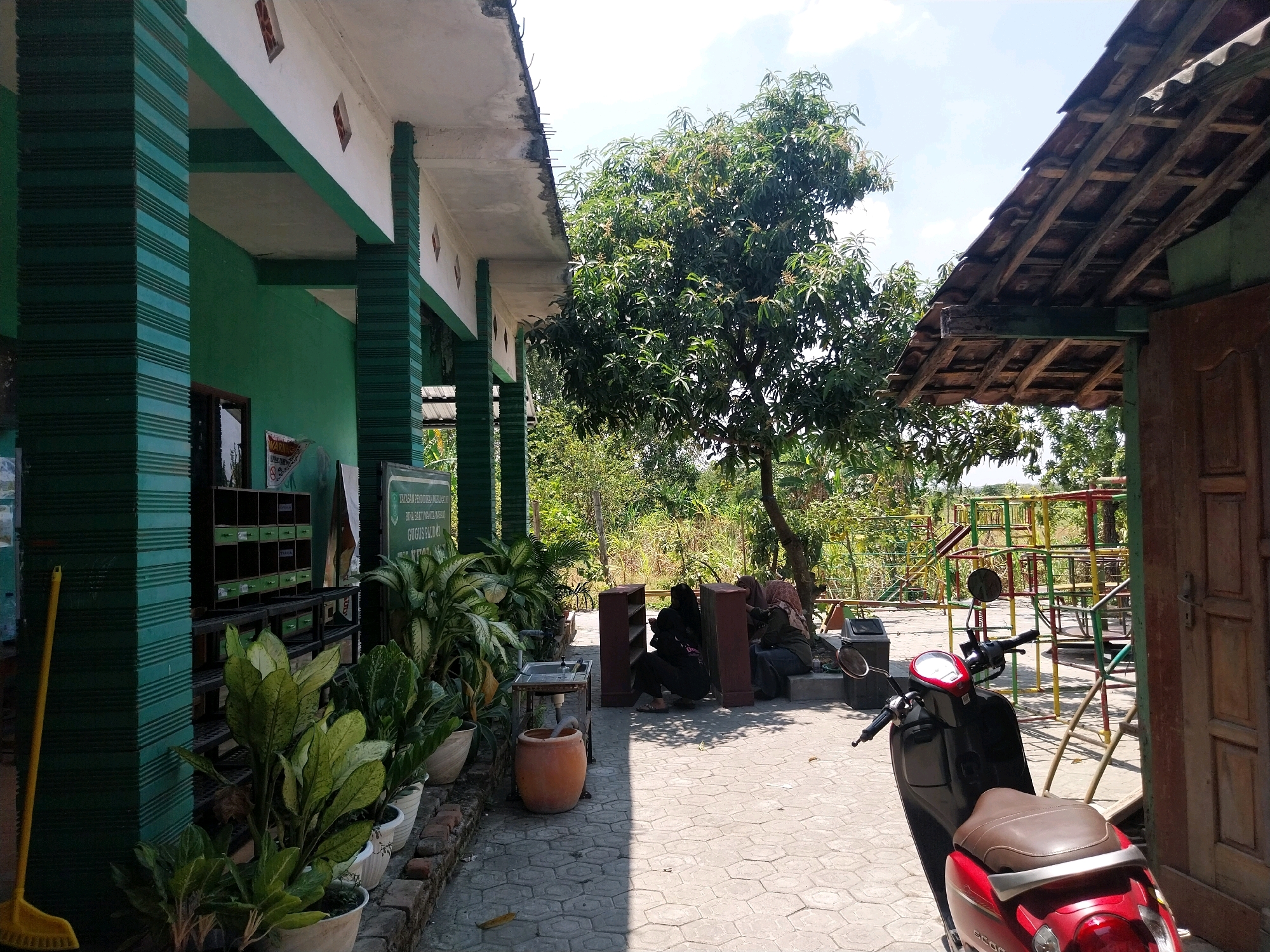 The image depicts an outdoor scene at a school in Campur, Indonesia, likely during the day. In the foreground, a red and black scooter is parked on a patterned paved area. To the left, a green building with white trim and dark green columns is visible, with potted plants in front of it. A yellow broom leans against one of the columns.
In the middle ground, several people are seated on the ground near dark brown shelves. Beyond them, a large green tree dominates the view, with a playground structure visible further in the background. The ground is paved with interlocking hexagonal stones. The sky is bright and cloudy. The overall impression is of a functional, somewhat weathered outdoor space at a school.