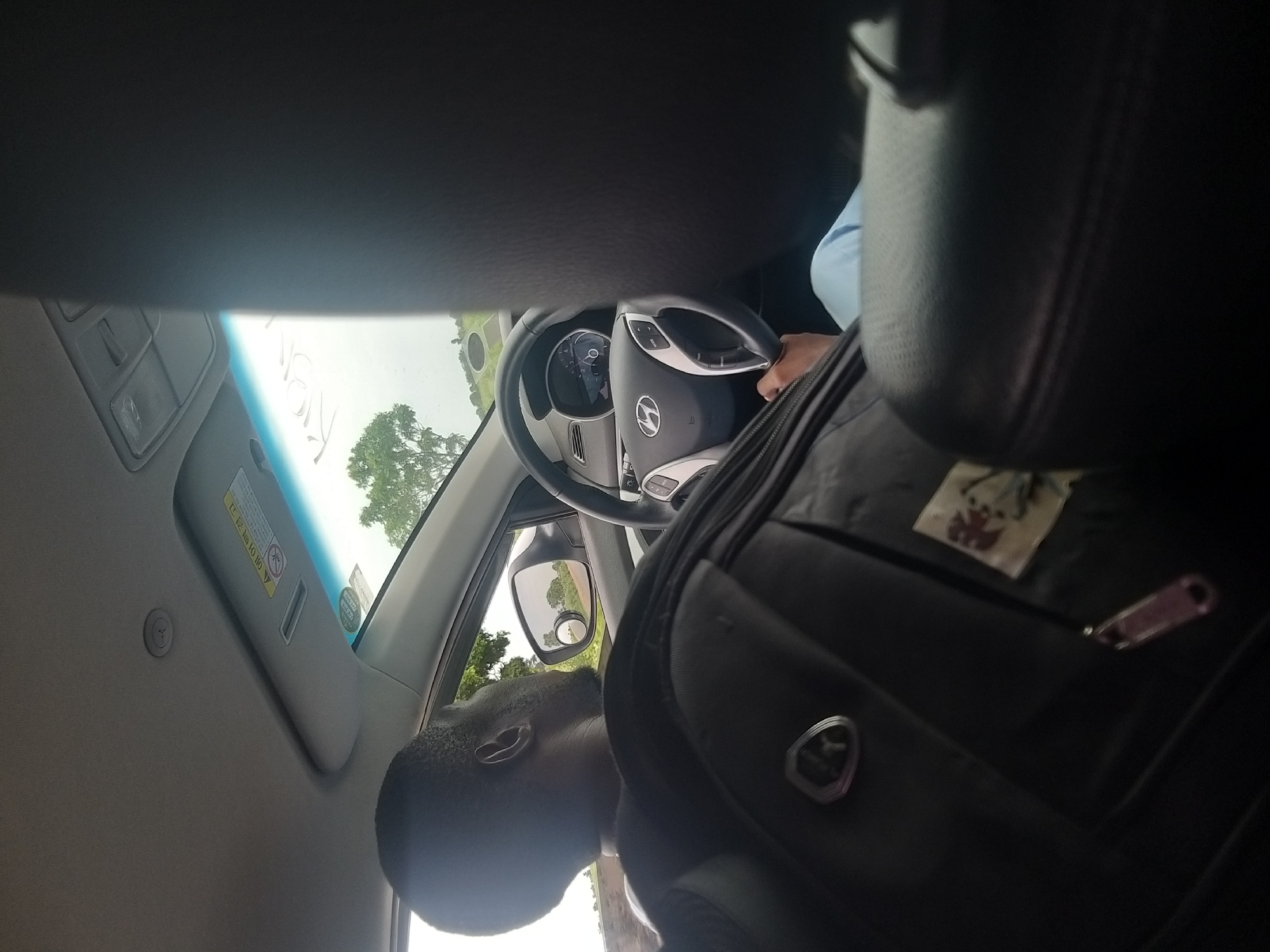The image shows the interior of a vehicle, viewed from the rear passenger seat. A man is visible from the shoulders up, looking out the side window. He has dark skin and short, dark hair. A large black bag is placed on his lap and extends forward, partially obscuring the driver's side of the car.
The driver's side of the car is more visible, with the steering wheel, dashboard, and speedometer clearly seen. The steering wheel has a silver Hyundai logo in the center. The dashboard displays various indicators. A hand, presumably the driver's, is resting on the steering wheel. The person appears to be wearing light blue pants.
Outside the car, visible through the windows, is a landscape with trees and what appears to be a road. A side mirror reflects a similar landscape. The sky is overcast. A sun visor is visible on the passenger side of the windshield, with some text and a warning symbol. The overall impression is that the vehicle is in motion.
While the image depicts the interior of a car, no specific elements within the image definitively place it in Kafarati, Nigeria. The scene is generic for a car interior in many locations.