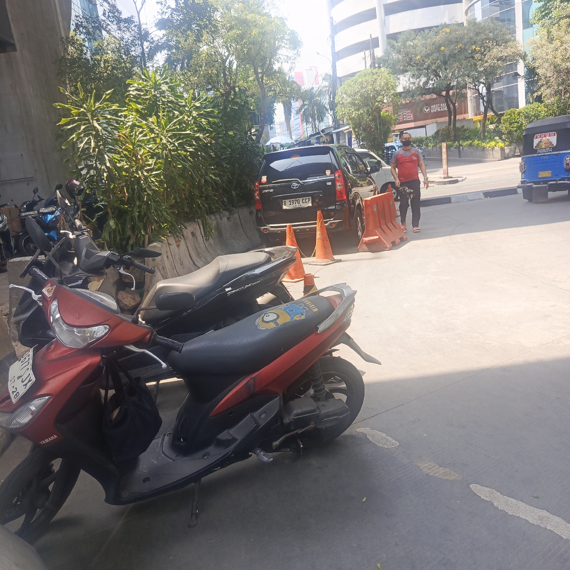 The image depicts an outdoor scene in South Jakarta, Indonesia, likely a parking area or street adjacent to buildings and lush vegetation. In the foreground, a red and black scooter is parked, with a black bag hanging from its handlebars. Behind it, another black scooter is visible. Further back, a black SUV with license plate "B 1970 CCP" is parked, flanked by orange traffic cones. To the right, a blue auto-rickshaw is parked. A man wearing a red shirt, dark pants, and a face mask is walking away from the camera, carrying a bag. In the background, modern buildings with glass facades rise above a row of trees. The ground is paved concrete with white painted lines and some darker stains. The overall lighting suggests a sunny day.