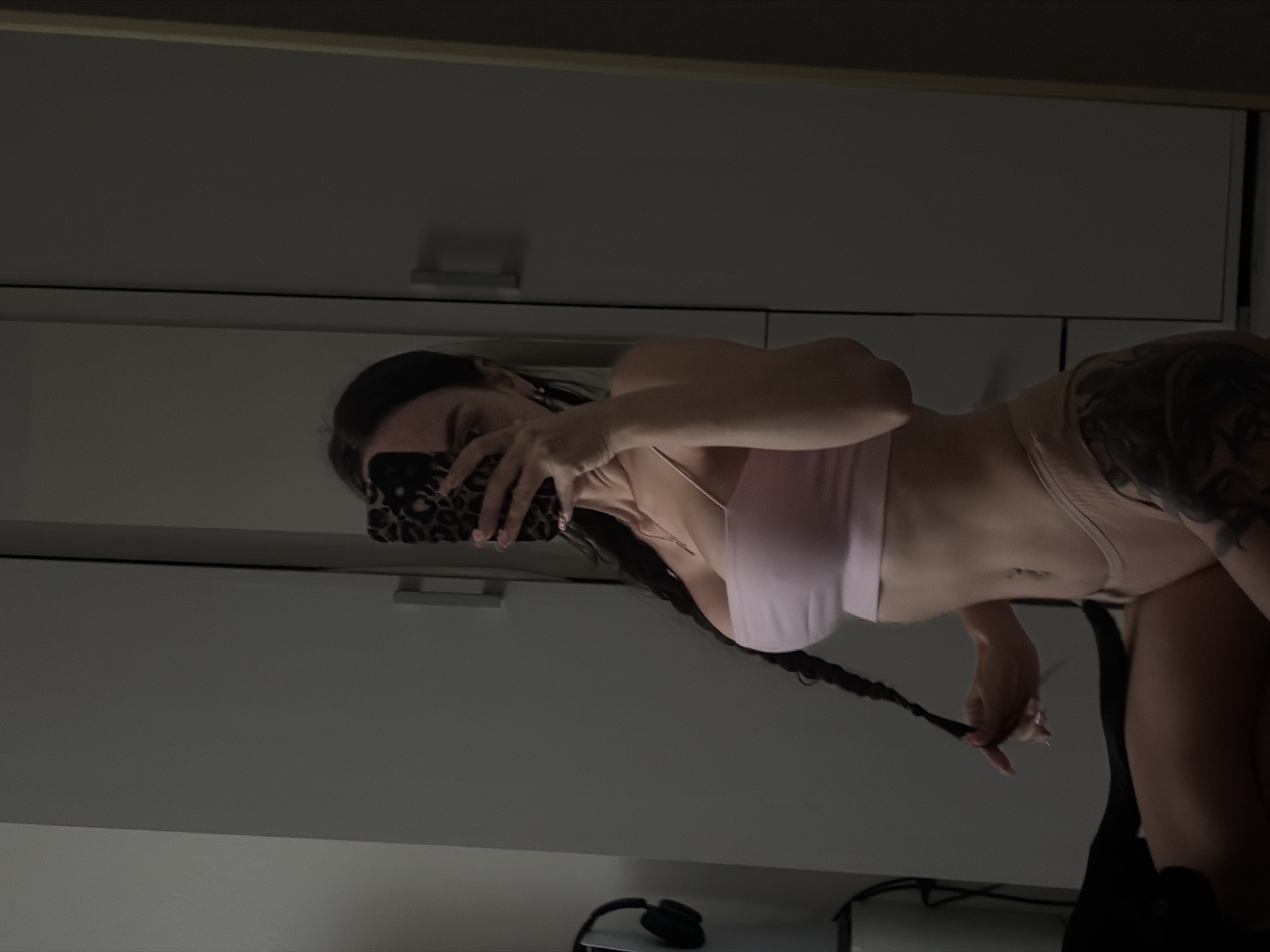 A woman with dark hair styled in a braid is taking a selfie in a room. She is wearing a pink crop top and beige underwear. The background consists of a closet and a mirror reflecting the scene. There is a pair of headphones on a stand to the left of the frame. The image is dark and appears to have been taken indoors. The location is not specified in the image or the prompt, therefore, it is not possible to determine if it is in Novosaratovka, Russia.