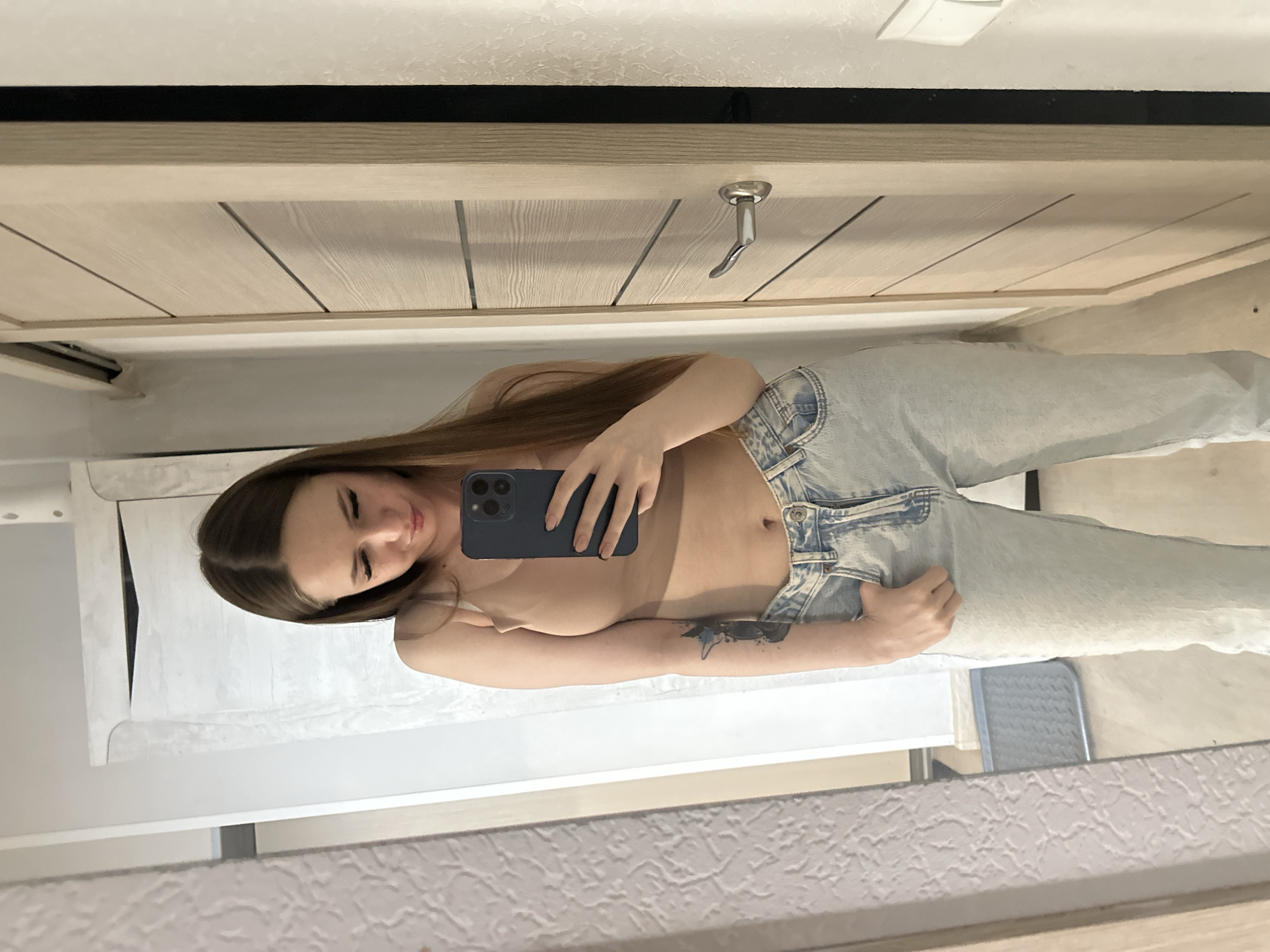 A woman is taking a selfie in a mirror. The woman has long brown hair and is wearing jeans and a beige top. She is holding a blue smartphone in her right hand and looking at the screen. The mirror is reflecting the room behind her, which appears to be a bathroom with a light-colored door and a white towel rack. The city is unknown.