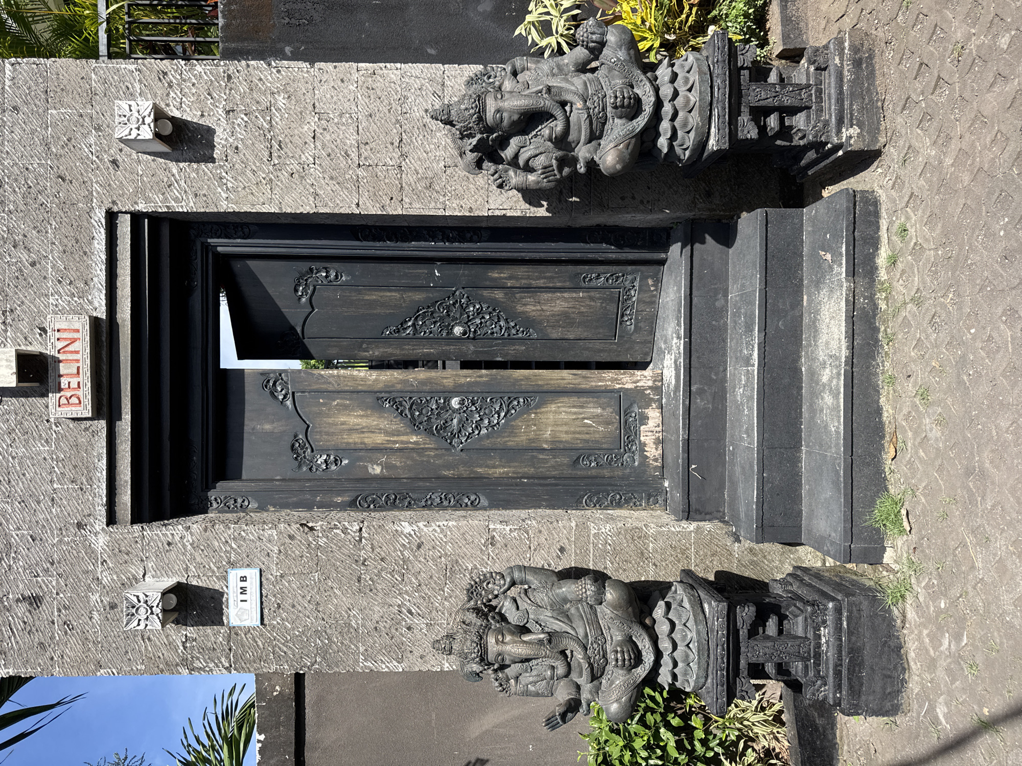 The image shows a stone entranceway with a dark, ornately carved wooden door slightly ajar. Two dark stone sculptures of what appear to be deities flank the entrance on either side. Above the door, there is a small stone cube attached to the wall. To the left of the door, a vertical sign reads "BELINI," while to the right, a square sign reads "IMB." The wall is made of diagonally striated stone. There are steps leading up to the entrance, and the ground surrounding the entrance is paved with stone tiles. Foliage is visible near the sculptures, including green plants and yellow-leaved plants. A palm tree and blue sky are visible in the bottom left corner. The photograph appears to be taken in Kuta Selatan, Indonesia, indicated by the name of the area on the sign.