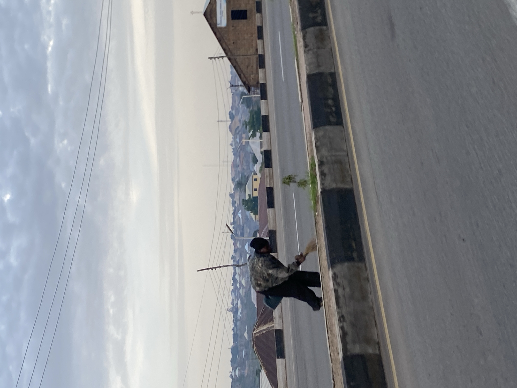 In Jos, Nigeria, a person in a camouflage jacket and dark pants sweeps the side of a road with a broom. The road has a concrete divider with black and white stripes. In the background, houses with red roofs are visible, along with a brown building that appears to be a church, indicated by a cross on its roof. The sky is overcast with clouds, and power lines stretch across the scene.