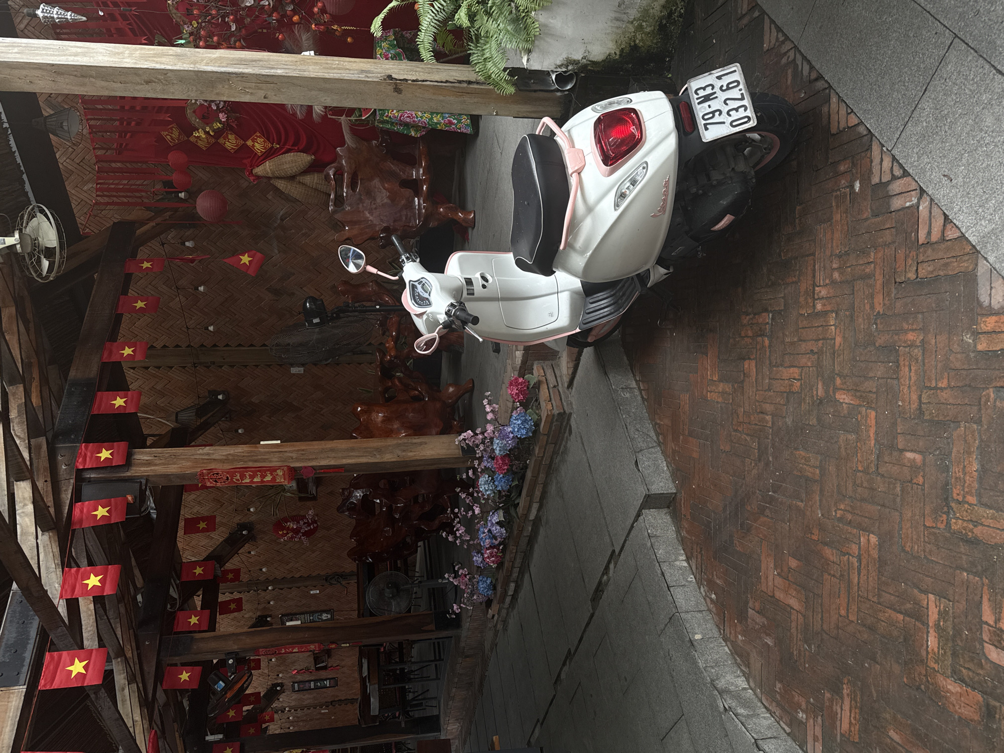 The image depicts a scooter parked in Bac Nha Trang Ward, Vietnam. The scooter, a white and pink Vespa, has a license plate reading "79-N3 032.61". It is parked on a brick and concrete walkway next to a building. The building is decorated with Vietnamese flags and ornate wooden furniture. There are also plants and flowers visible. The scene is well-lit with natural light.