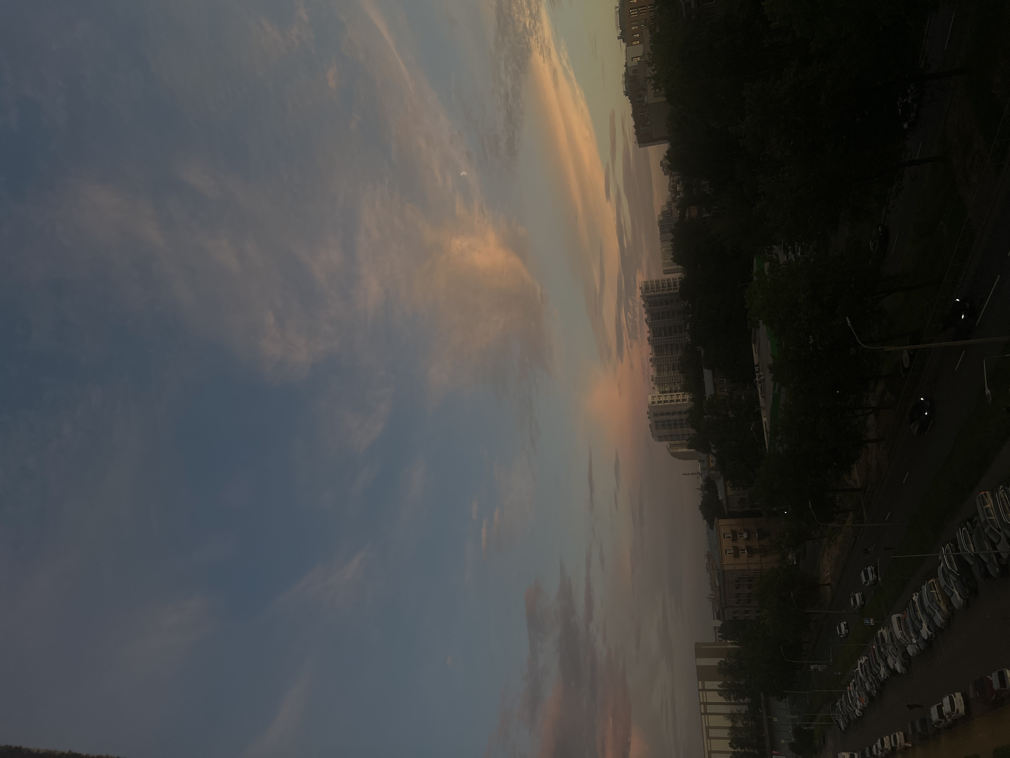 The image depicts a dusk sky over Petro-Slavyanka, Russia. The sky displays a gradient of blue and soft pink and orange hues, with scattered wispy clouds. A sliver of a crescent moon is visible in the upper right quadrant of the sky. Below the sky, a cityscape is silhouetted against the fading light. Numerous apartment buildings of varying heights are visible, interspersed with dark green trees. In the foreground, a street is lined with parked cars and a few streetlights are illuminated. There are no people clearly visible in the scene. The overall impression is of a residential area in a city during twilight.