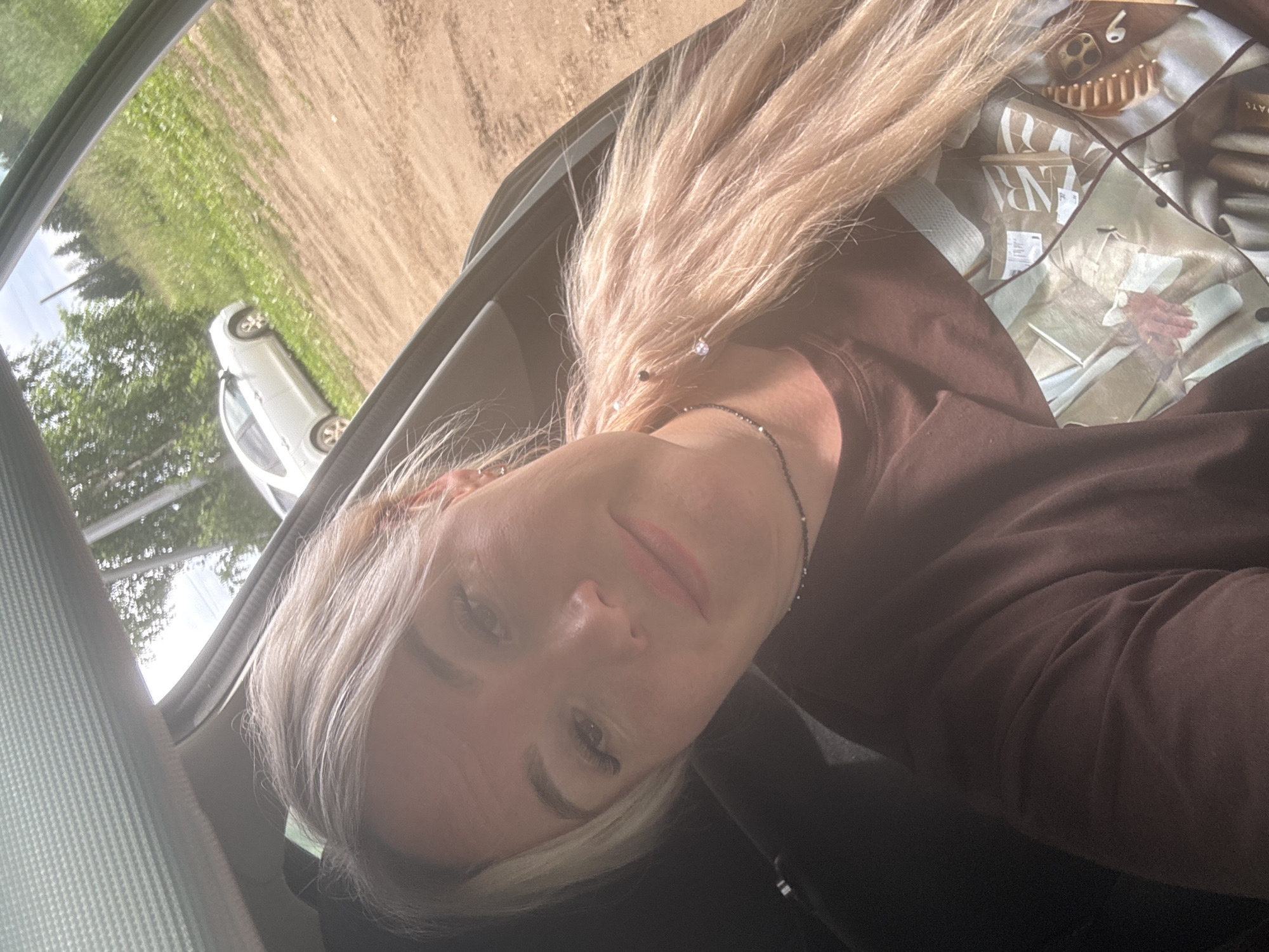 A woman with blonde hair is taking a selfie inside a car. The car appears to be parked on a dirt road with green vegetation and trees visible outside the windows. A white car is parked in the background. The woman is wearing a brown hoodie and a necklace. She has makeup on and her hair is styled down. The photo has a slightly overexposed, bright, daytime feel, suggesting it was taken on a sunny day. The background inside the car includes a patterned fabric, possibly a seat cover or bag, with some text that is partially visible and appears to include "ZARA". The woman's expression is relaxed and pleasant, hinting at a casual moment. The overall scene suggests a spontaneous snapshot taken during a car ride or stop.