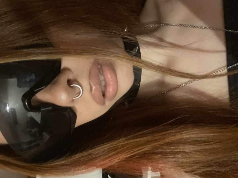 A head-and-upper-torso portrait features an individual with long, reddish-brown hair, displaying a distinct lighter red streak on the viewer's left side. The person is wearing large, dark, reflective sunglasses that completely obscure their eyes. A silver-colored hoop septum piercing is visible in the nose. The lips are parted slightly, revealing upper teeth, and appear full with a glossy finish. Around the neck, a black choker adorned with silver-colored studs is worn, beneath which a thin silver-colored chain necklace hangs. The visible clothing on the upper body is dark. The background is out of focus and indistinct, indicating an indoor environment. The location is Unknown, Unknown.