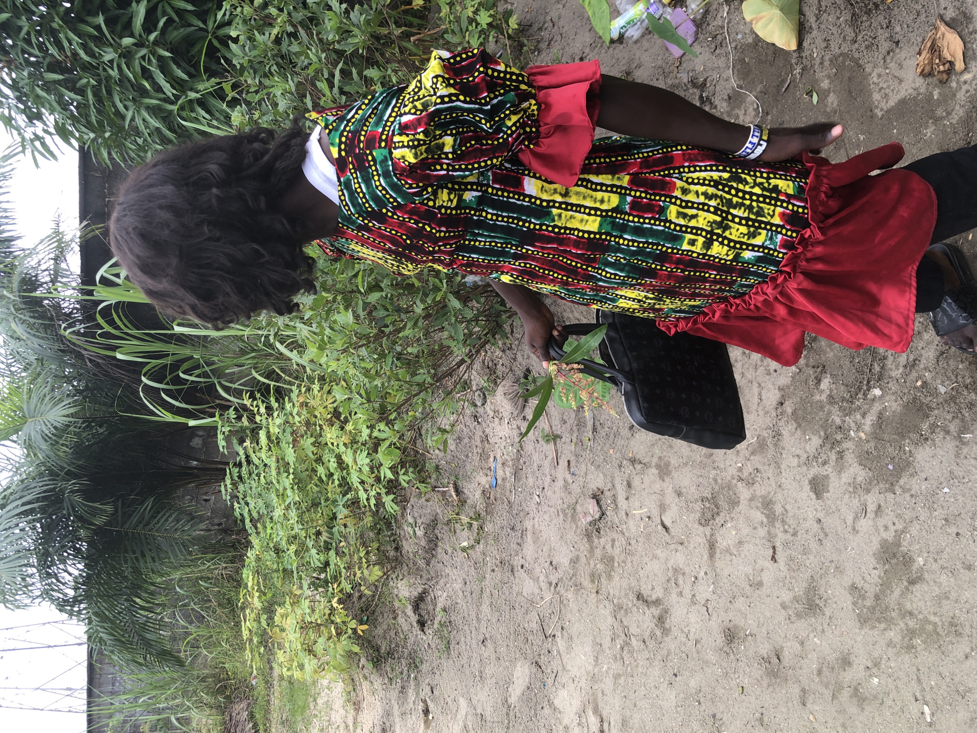 A young person with dark, curly hair stands in a dirt area. They are wearing a colorful, patterned dress with red ruffled trim and a white undershirt. The dress is made of fabric with vertical stripes of green, yellow, and red, with black and white dots forming patterns within the stripes. On their left arm, they wear a white bracelet. In their right hand, they hold a black handbag with a textured pattern. The ground is composed of sand and dirt, with some scattered debris. Behind the person is a dense growth of green plants and palm trees, with a gray brick wall visible in the background. The overall environment appears to be an outdoor setting in a rural or semi-rural area, potentially on the outskirts of Ado, Nigeria, suggested by the presence of lush vegetation and unpaved ground.