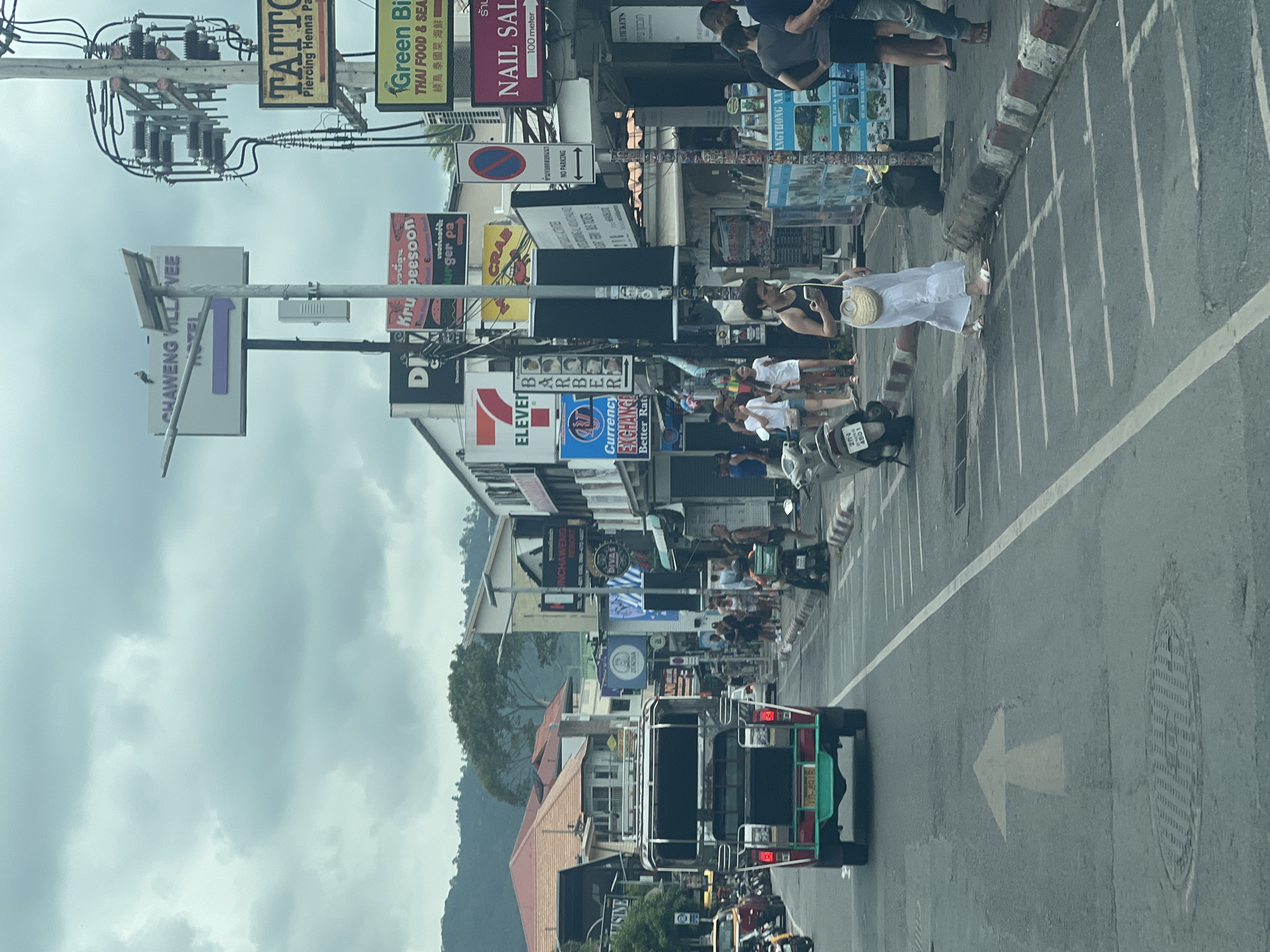 The image depicts a street scene in Chaweng, Ko Samui District, Thailand. A red and green tuk-tuk is visible in the foreground, moving away from the viewer. A yellow arrow is painted on the asphalt road, indicating a direction of travel. The street is lined with various shops and signs, including a "CHAWENG VILLAGE HOTEL", "TATTOO", "Green Big", "NAIL SALON", and "7-ELEVEN". Several people are walking on the sidewalk and crossing the street. The sky is overcast, suggesting a cloudy day. The buildings are mostly two to three stories high and display a variety of architectural styles. A utility pole with numerous wires and electrical equipment is prominent on the right side of the image. Several motorcycles are parked along the street. The environment appears to be a bustling commercial area.