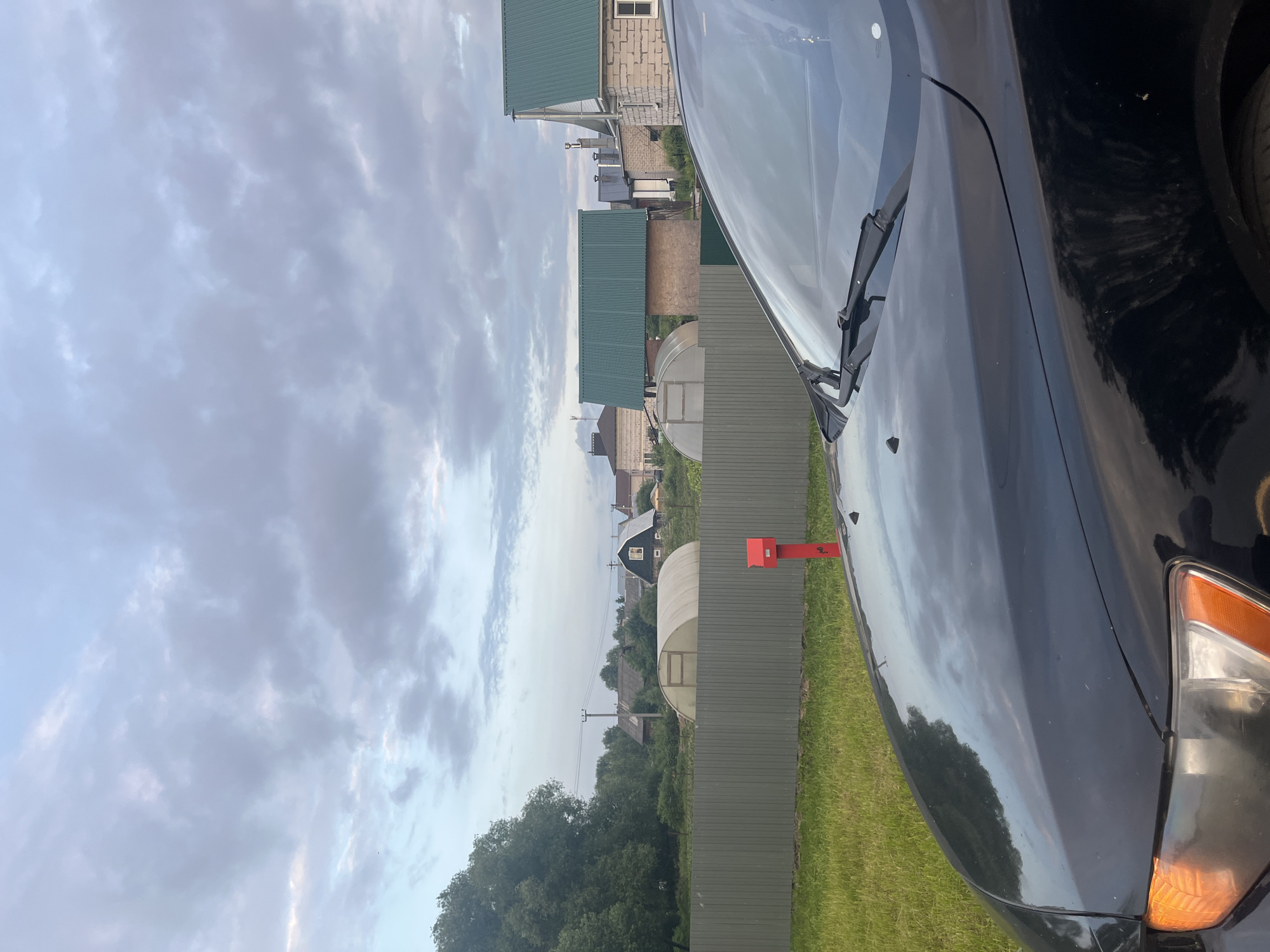 This media file captures a rural scene in Nikolskoye, Russia, likely during the late afternoon or early evening given the soft, diffused light and cloudy sky. The image is taken from the perspective of a person standing near a black car, which occupies the right side of the frame. The car's hood and headlight are visible, reflecting the cloudy sky and surrounding environment.
The scene is characterized by a collection of buildings with green corrugated roofs, typical of dachas or rural homes. Several of these structures are visible in the background, interspersed with trees and vegetation. A greenhouse with a transparent roof is also present. A red mailbox stands prominently on the left side of the frame, near a green metal fence that separates the yard from the buildings and further greenery.
The weather appears overcast with dramatic, textured clouds filling the sky. The ground is covered in green grass, suggesting a temperate climate and possibly summer or late spring. There is no visible text in the image. The overall mood is peaceful and tranquil, evoking a sense of rural solitude. The composition, with the car in the foreground and the village in the background, provides a glimpse into the daily life or landscape of this Russian locality.