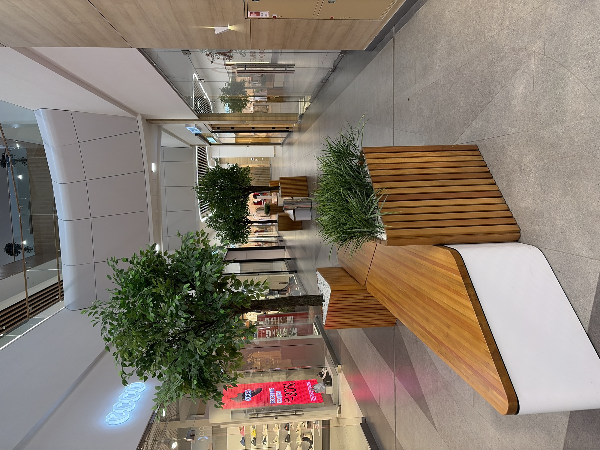 This image depicts the interior of a brightly lit, modern shopping mall corridor. The location is the Talisman Shopping and Entertainment Center (ТРЦ Талисман) in Izhevsk, Udmurt Republic, Russia.
The scene is viewed from a slightly low angle, looking down a wide, empty walkway. In the immediate foreground on the left is a distinctive, low-profile bench with a white base and a polished wooden top, shaped with a rounded, triangular end. Adjacent to this bench on the right is a tall rectangular planter, constructed from vertical wooden slats, containing vibrant green artificial grass and white decorative stones.
Further into the midground, the corridor is punctuated by several large, artificial trees housed in similar wooden-slatted planters filled with white stones. These trees are arranged in a linear fashion along the left side of the walkway, separating the main path from the storefronts.
On the left side of the corridor, a prominent "ECCO" shoe store is visible, identified by a bright blue neon sign spelling out the brand name above its entrance. A large, vertical red banner inside the store window advertises "ВЕСЕННИЕ СКИДКИ ДО -30%" (Spring Discounts Up To -30%), accompanied by the ECCO logo. Various shoes are displayed on shelves within the store.
On the right side of the corridor, another brand, "GEOX," is visible on a store facade with golden accents. Further down, reflections on glass storefronts hint at other brands, including "MUSTANG." The right wall is clad with light brown wood panels and features recessed lighting fixtures. A beige utility cabinet, likely a fire safety cabinet, with labels "TK-42" and "10-42" is mounted on this wall.
The floor is tiled with large, light gray square tiles, incorporating subtle, lighter gray geometric patterns. The ceiling is high and features a contemporary design with white panels and integrated strip lighting. An upper level of the mall is visible, characterized by a curved white structure and transparent glass railings, reflecting the overhead lighting. No people are present in the scene.