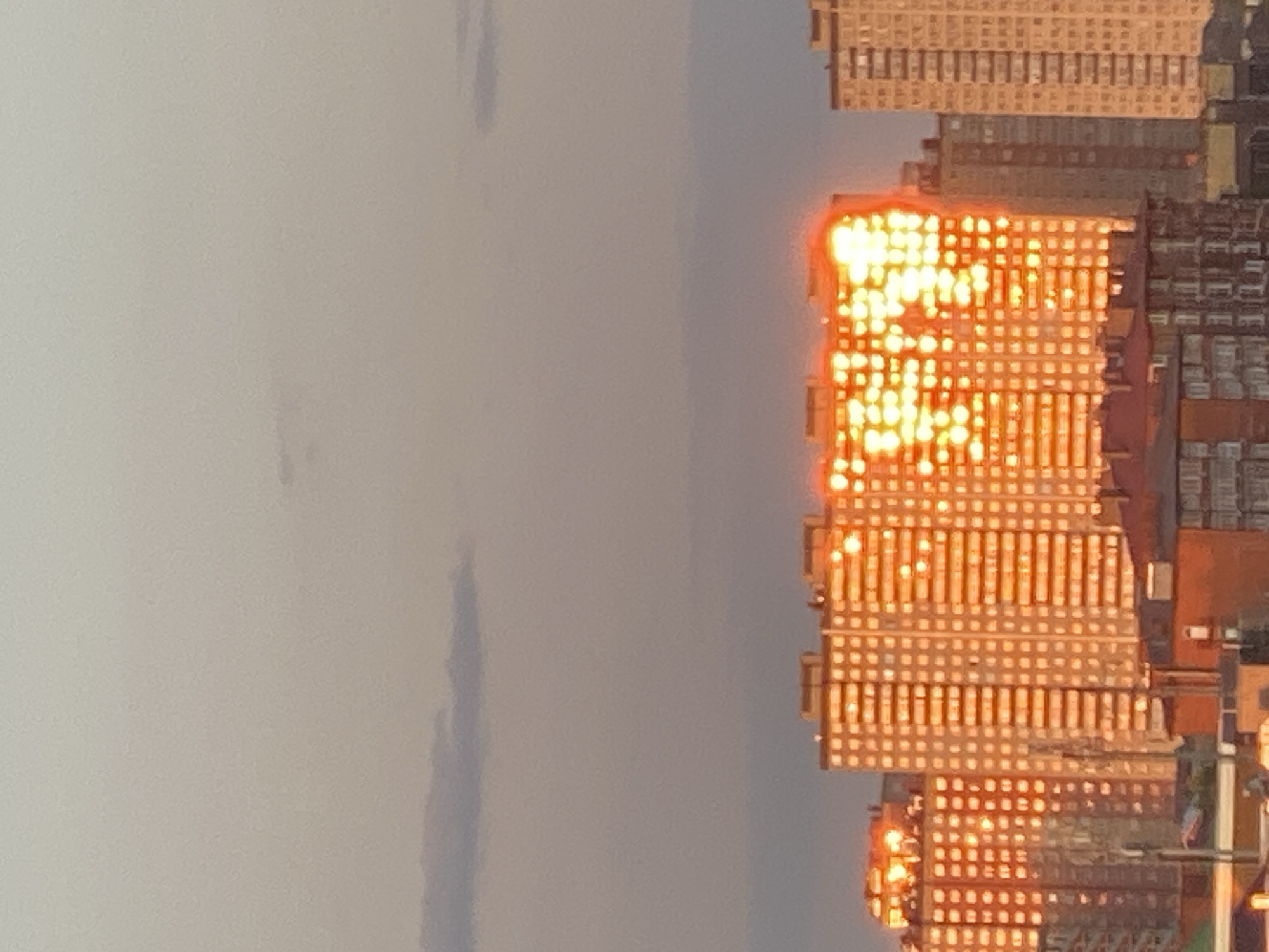 This vertically oriented image captures an urban landscape, likely in Krasnodar, Russia, featuring several high-rise buildings illuminated by intense, golden-orange light. The buildings, which occupy the right side of the frame, display a grid-like pattern of windows, many of which are brightly reflecting what appears to be a low-angled sun. This reflection creates a shimmering, almost glowing effect across the facades, particularly on the prominent building in the mid-right.
The sky, filling the left side of the image, is a uniform, light gray, indicating overcast or hazy weather conditions. This muted sky provides a strong contrast to the vibrant, warm light reflecting off the buildings. The distinct glow on the buildings suggests either sunrise or sunset. Details of the buildings' architecture are visible, showing multiple stories and structural elements, but are somewhat overshadowed by the powerful glare. No people or discernible text are visible in the scene. The overall atmosphere conveys an urban environment at a specific, striking time of day.