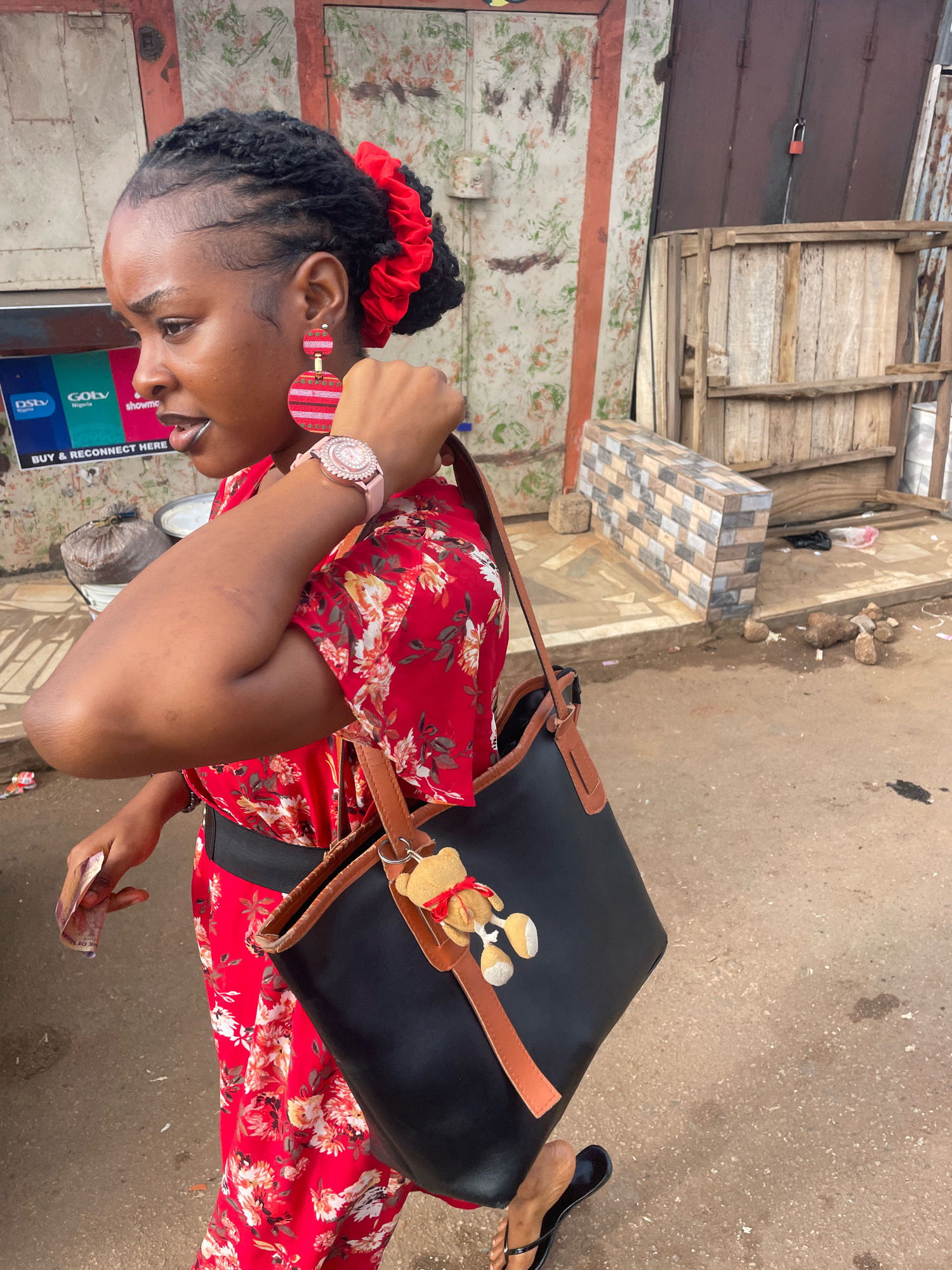 A full-shot, eye-level, outdoor, medium-wide photograph captures a young Black woman in Gura Top, Nigeria. She is looking to her left with her mouth slightly open, as if speaking. The woman has dark, braided hair styled into a bun with a large red scrunchie. She is wearing large, circular, pink and red striped earrings and a pink watch with a sparkly face. Her red, short-sleeved dress features a white and orange floral pattern. She is carrying a black tote bag with tan leather straps over her right shoulder. A small, brown teddy bear charm is attached to the bag. In her left hand, she is holding a rolled-up bill. The background shows a weathered, green and white painted wall with a red stripe, a dark metal door with a padlock, and a wooden crate. To the left of the woman, there is a white poster with logos for DStv and GOtv. The ground is dirt and concrete with scattered rocks and debris.