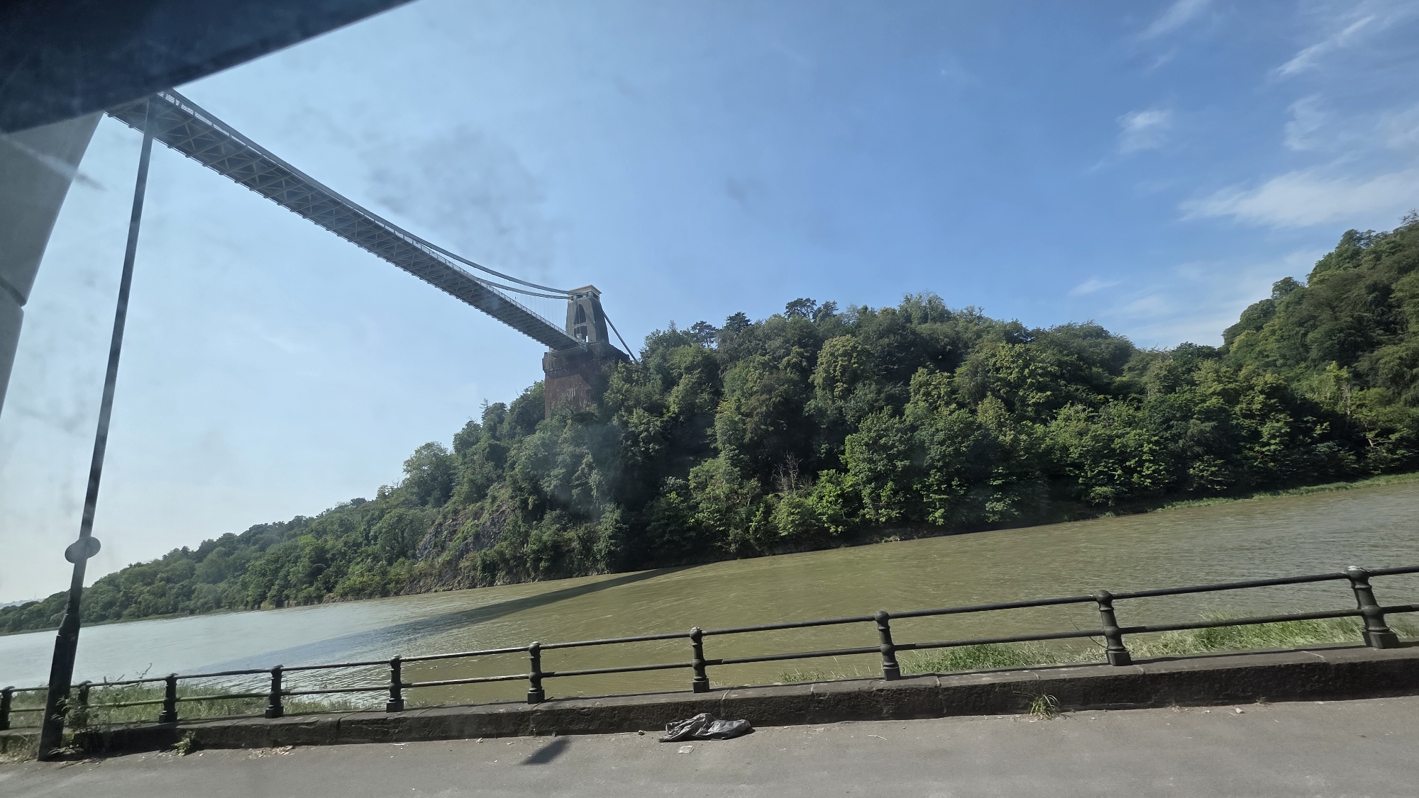 The image shows the Clifton Suspension Bridge spanning the Avon Gorge in Bristol, United Kingdom. The bridge, a famous landmark, is seen from a low angle, with its iconic towers and suspension cables silhouetted against a clear blue sky dotted with wispy clouds. The gorge is densely wooded, with lush green trees covering the steep cliffs. A wide, muddy river flows below the bridge, reflecting the sky and trees. In the foreground, a dark metal railing lines a paved walkway or road, separating it from the river. A single dark plastic bag is lying on the ground near the railing. The overall impression is one of natural beauty and impressive engineering.