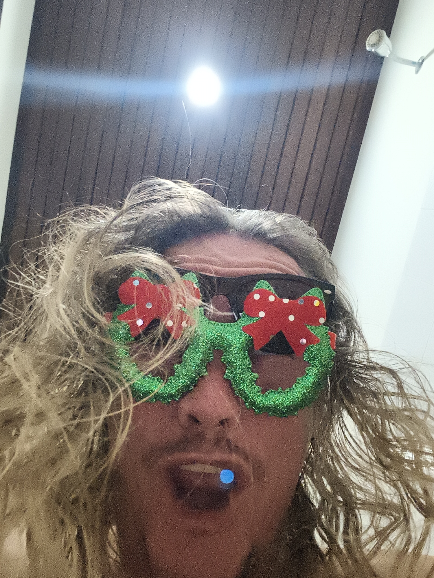 A low-angle close-up shot shows a person wearing Christmas-themed glasses with a green wreath frame and red bow accents. The person has light-colored, wavy hair, sunglasses underneath the festive glasses, and is sticking their tongue out with a small blue object on it. The background shows a dark wood-paneled ceiling and a portion of a white wall with a light fixture. The location is บ้านบางรักษ์, Thailand.