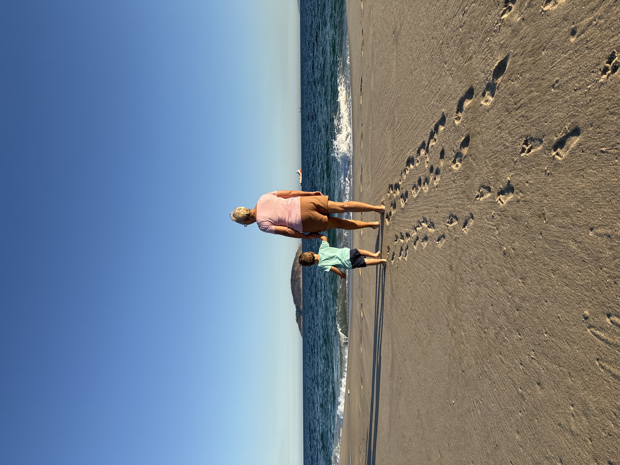 The image shows a woman and a young boy walking on a sandy beach in Niterói, Brazil. The woman, seen from behind, wears a pink t-shirt and brown shorts, with blonde hair and glasses. The boy, also seen from behind, is wearing a turquoise t-shirt and dark shorts. They are walking towards the ocean, leaving footprints in the sand. The sea is blue with gentle waves, and in the distance, an island with a green top is visible. A cargo ship is also seen on the horizon. The sky is clear and blue. The scene captures a moment of a leisurely walk along the coastline.