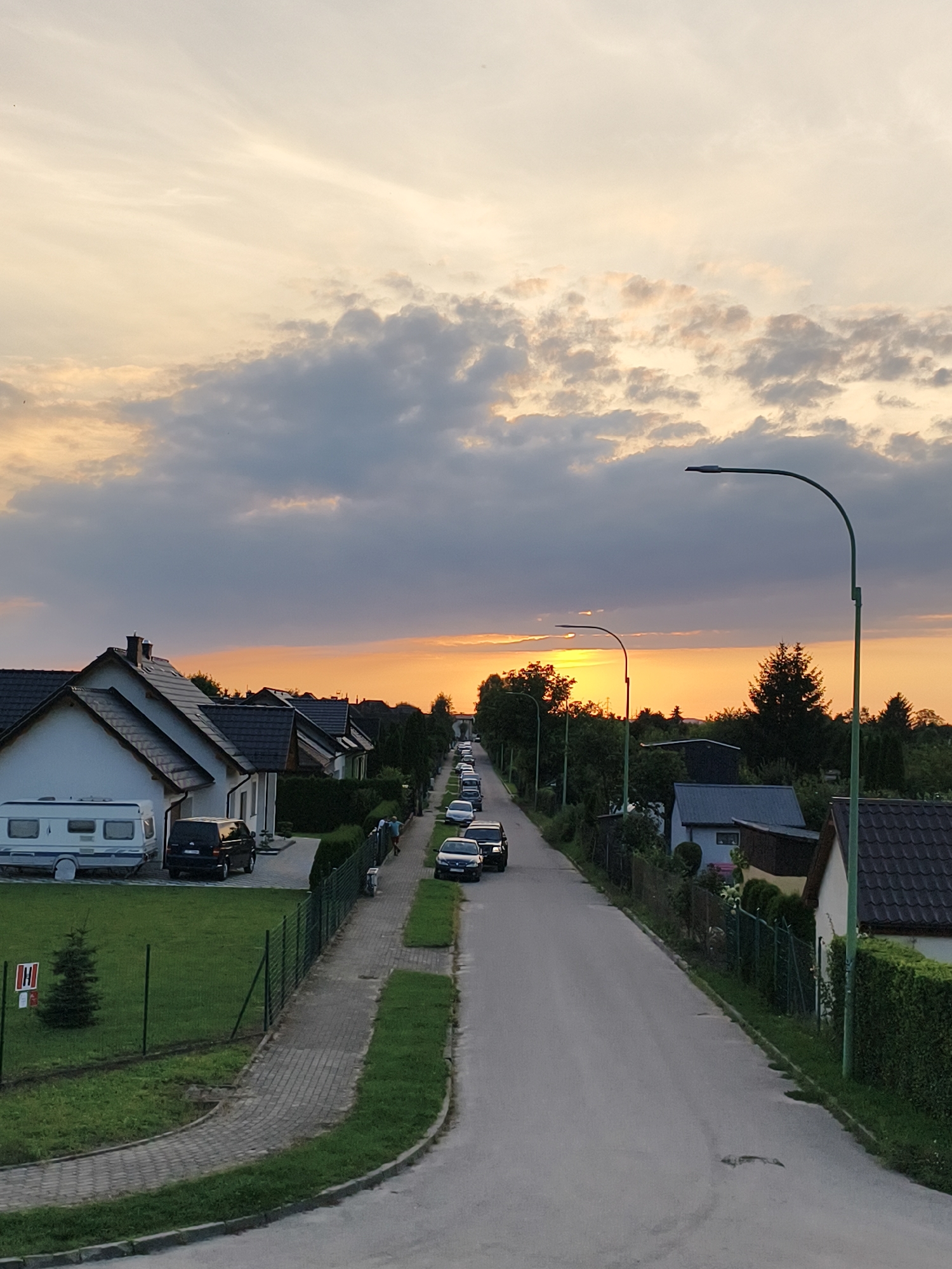 The image shows a suburban street at sunset in Tczew, Poland. The sky is filled with dramatic clouds, with the sun setting behind a line of trees. On the left side of the street are several houses with dark roofs, and a white camper van. A black minivan is parked in a driveway. A paved sidewalk runs alongside the street, bordered by a green lawn and a fence. On the right side of the street are more houses, also with dark roofs, and more trees. Several cars are parked along the street and on the right side of the road. A street light with a green pole and a modern lamp is visible on the right side of the image, and another similar lamp is visible further down the street on the left. The overall impression is one of a quiet residential neighborhood at the end of the day.