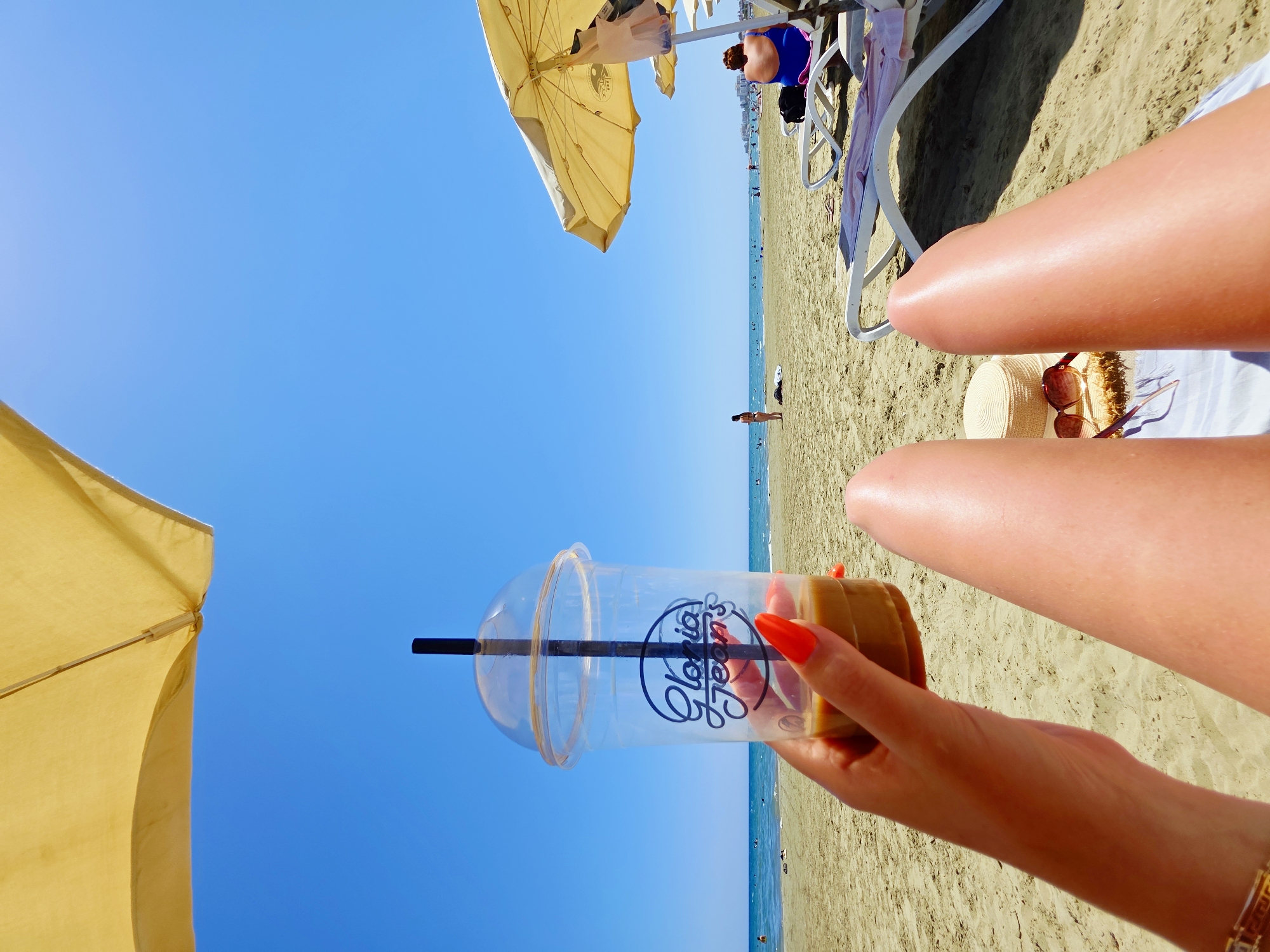 A first-person perspective shot shows a person's hand holding a clear plastic cup with a black straw and a brown liquid, branded with "Gloria Jean's," while sitting on a beach. The person's legs are visible in the foreground, with a straw hat and sunglasses on the sand next to them. In the background, the sea meets a sandy beach with several people scattered about, and a few umbrellas providing shade. The scene is set under a clear blue sky on a sunny day. The location is implied to be Larnaca, Cyprus, a popular tourist destination known for its beaches.