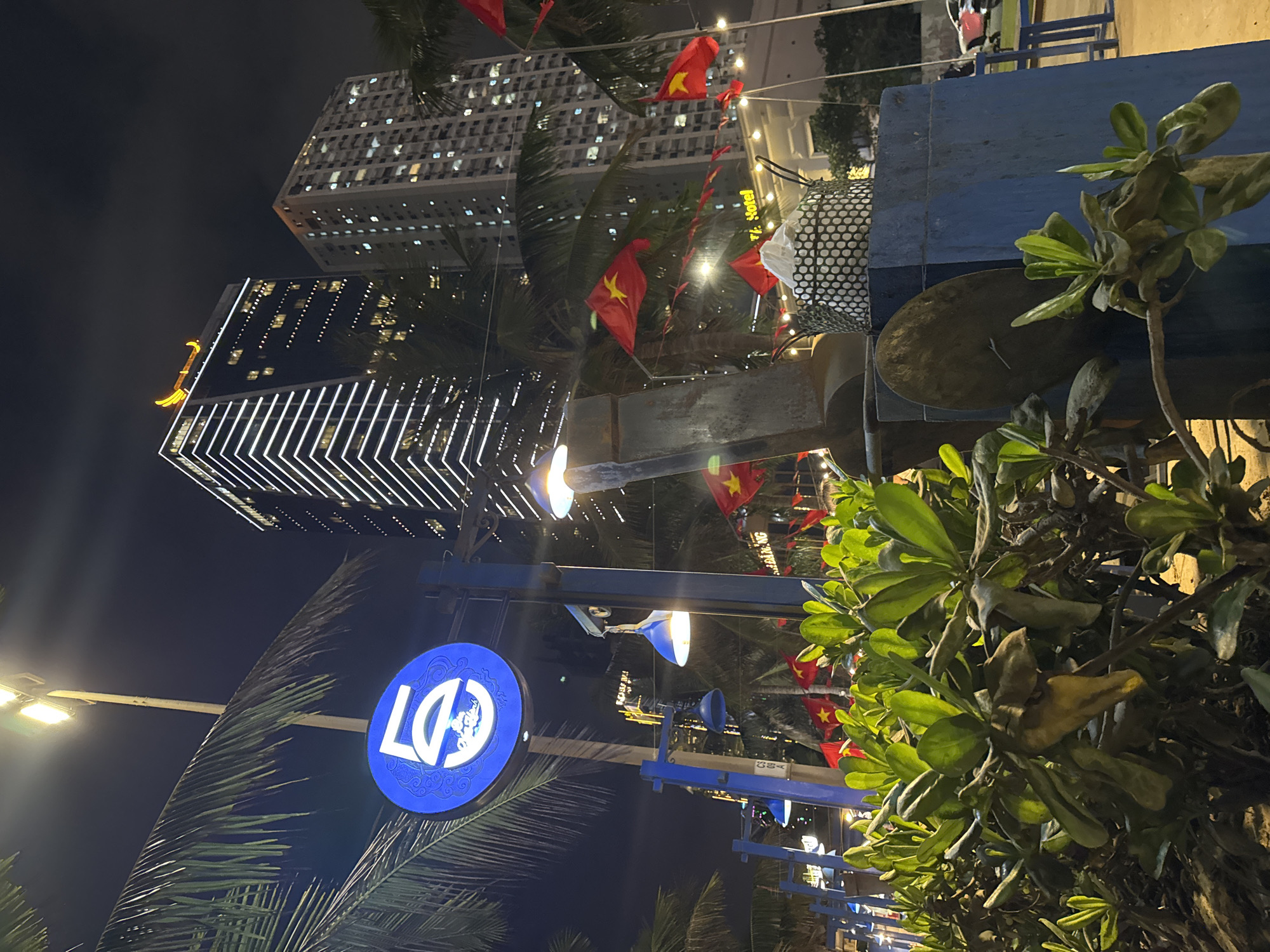 The image captures a vibrant night scene in Đà Nẵng, Vietnam, showcasing a blend of modern urban architecture and tropical greenery. In the background, two towering skyscrapers rise against a dark sky, their windows illuminated, with one featuring striking white vertical strip lights and a golden, curved embellishment at its apex.
The foreground is filled with lush green foliage, including discernible palm fronds, alongside blue structural elements that suggest outdoor seating or a decorated promenade. Numerous small Vietnamese flags (red with a yellow star) are strung as decorations throughout the scene, adding a festive touch. The area is brightly lit by streetlights and decorative string lights.
A prominent blue circular sign with white lettering displaying "LĐ" and a small graphic of a figure is visible on the left side. Faintly visible text on a distant building facade appears to read "T.K. Hotel." The overall atmosphere is lively and well-lit, indicative of a popular urban or tourist area at night, under clear weather conditions.