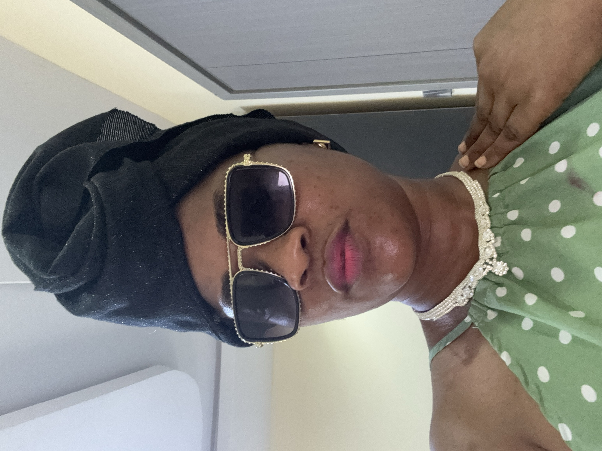 A woman wearing a black headscarf and sunglasses is shown from the chest up. She has a green and white polka dot dress on, with a jeweled necklace. She is in a room with light colored walls.