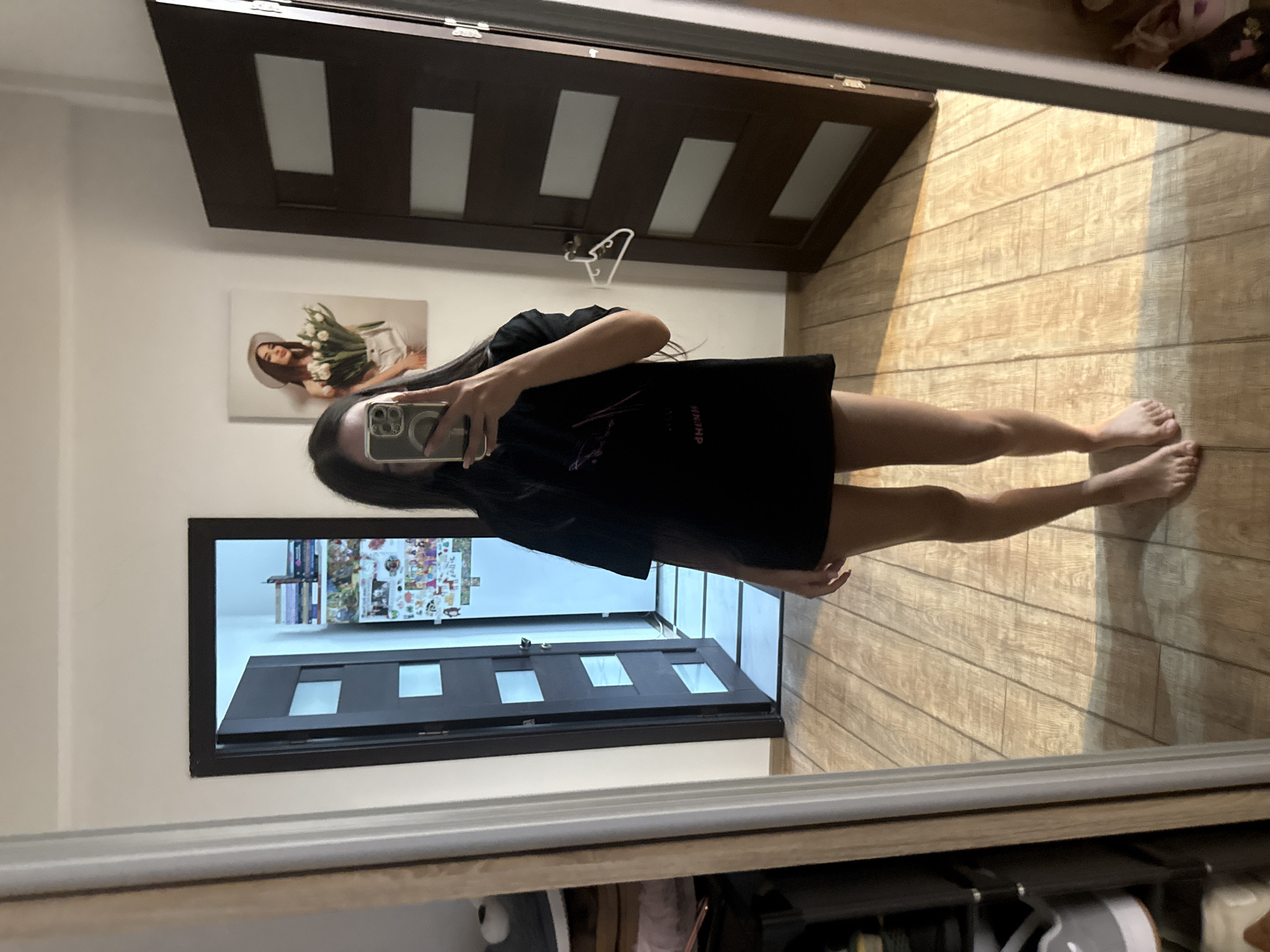A young woman with long dark hair is standing in a room, taking a mirror selfie. She is wearing a black t-shirt and no pants, with her bare legs visible. Her phone, with a clear case, is held up to take the picture. The background shows a wooden floor, two interior doors, and a framed picture of a woman holding tulips and wearing a hat on the wall. The overall scene appears to be in a residential setting, possibly an apartment or house, within the city of Chișinău, Moldova.