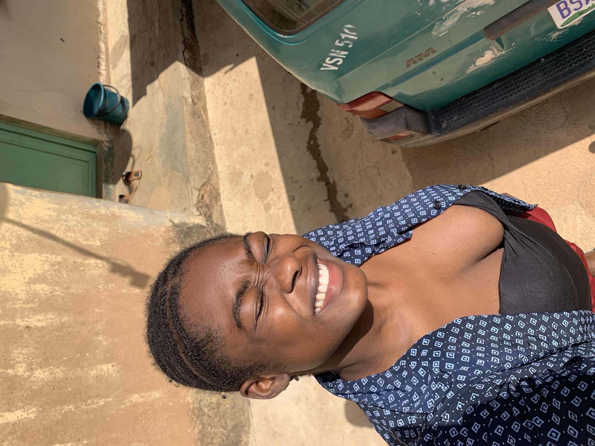 The image shows a smiling woman with dark hair and skin, wearing a dark blue patterned shirt. The photograph appears to have been taken outdoors. The background includes a green vehicle with a license plate partially visible, "VSN 510," and another plate starting with "BS". The location shows a light tan or beige wall or building exterior. There is a light blue bucket mounted on the wall, a green door, and the ground appears to be a rough, concrete or stone surface. Based on the query, the location is Rafin Par, Nigeria.