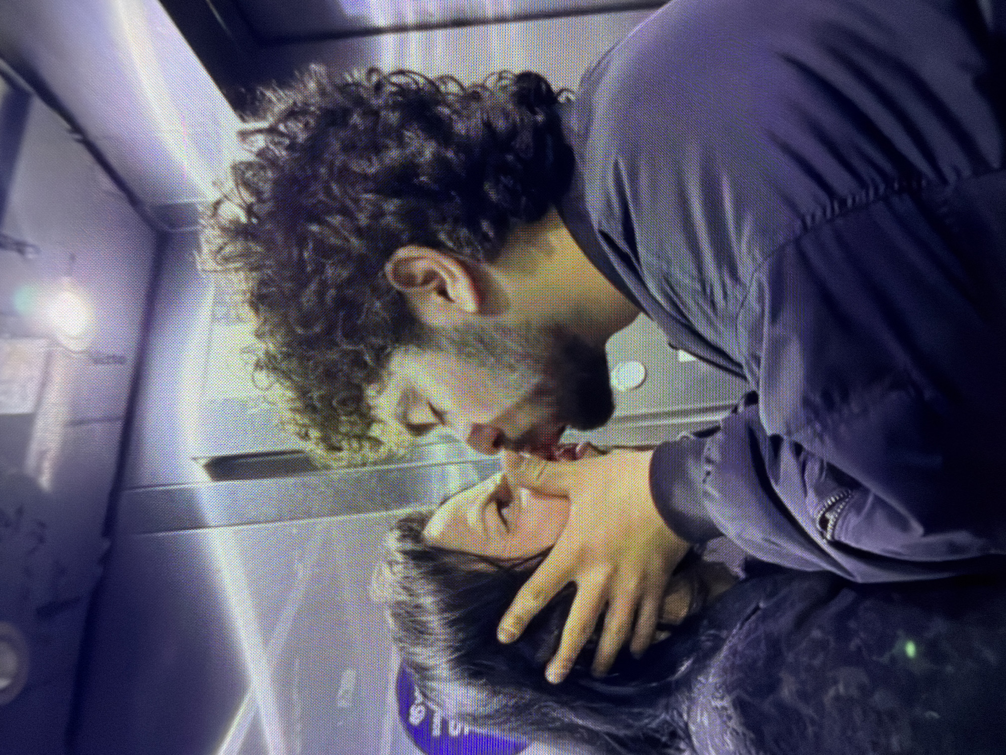 A man and a woman are kissing in an elevator. The man has curly hair, a beard, and is wearing a dark jacket. He is holding the woman's face with his hand. The woman has dark hair. The interior of the elevator is visible in the background. The location of the city is unknown.