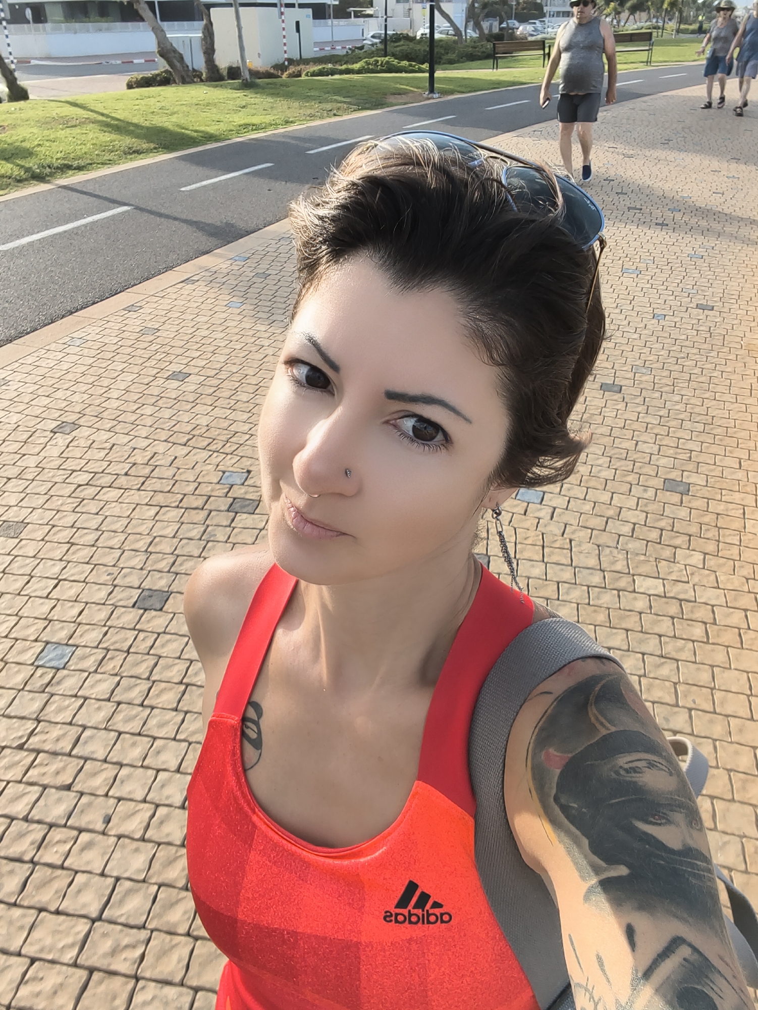 In a sun-drenched, paved walkway in Nahariyya, Israel, a woman with short, dark hair is taking a selfie. She is wearing a bright red athletic tank top with the Adidas logo visible on the front. A grey strap from a backpack is visible over her shoulder, and a large tattoo adorns her left arm. She has a nose ring and wears sunglasses pushed up onto her head.
In the background, a man in a grey tank top and dark shorts walks away from the camera, holding a phone. Further in the distance, another person in a light-colored hat and blue shorts is also walking along the path. The walkway is bordered by a grassy area and a road, with trees and buildings visible in the far background. The light suggests it is late afternoon or early evening.