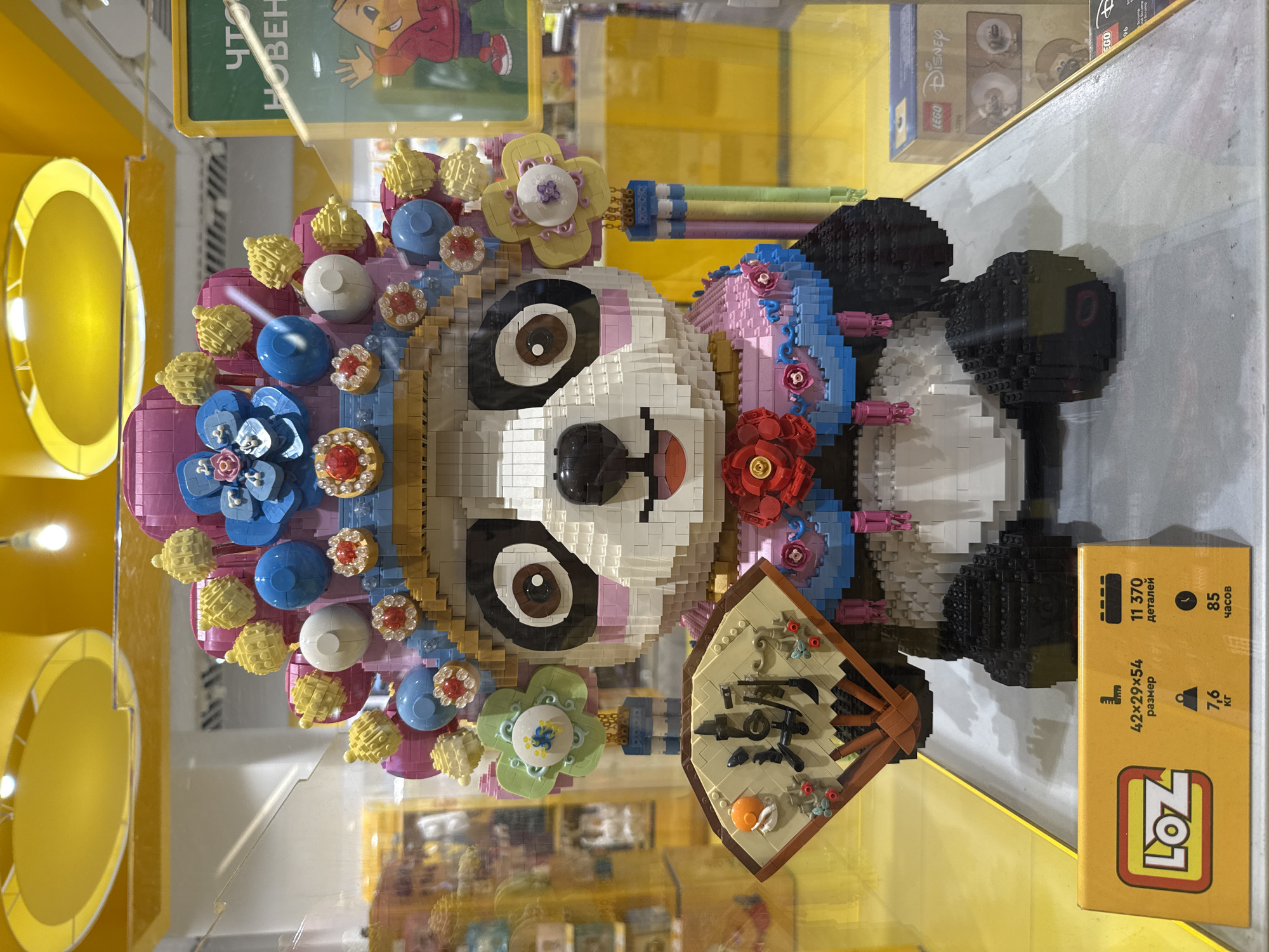 The image displays a large, intricately constructed Lego panda figurine dressed in traditional Chinese opera attire, complete with a headdress adorned with flowers and pearls, and holding a fan with Chinese characters. The panda is positioned in a retail display, likely a toy store, behind a glass enclosure. The background shows a brightly lit environment with yellow ceiling fixtures and shelves stocked with merchandise. A yellow price tag in the foreground provides product details, including "LOZ" branding, dimensions, weight, and assembly time. The reflection on the glass suggests the photo was taken in a public space. The retail environment, featuring toy displays and price tags, indicates this scene is located within a store. While the image shows a Lego display, its specific location in Yaroslavl, Russia, cannot be definitively determined from the visual information provided.