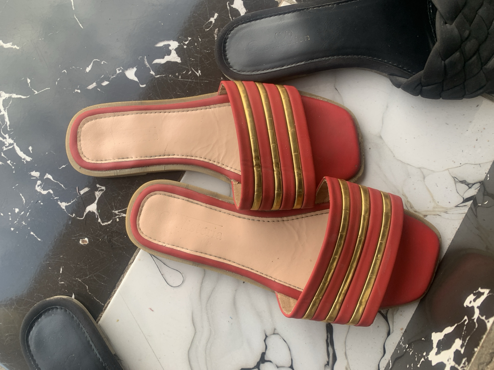A pair of red slide sandals with gold straps are displayed on a marble-patterned surface. To the left, a black sandal is partially visible, and to the right, another black sandal with a woven strap is also shown. The sandals appear to be indoors, possibly on a tiled floor or a table. There is no indication of the specific location within Jauro Yinu, Nigeria.