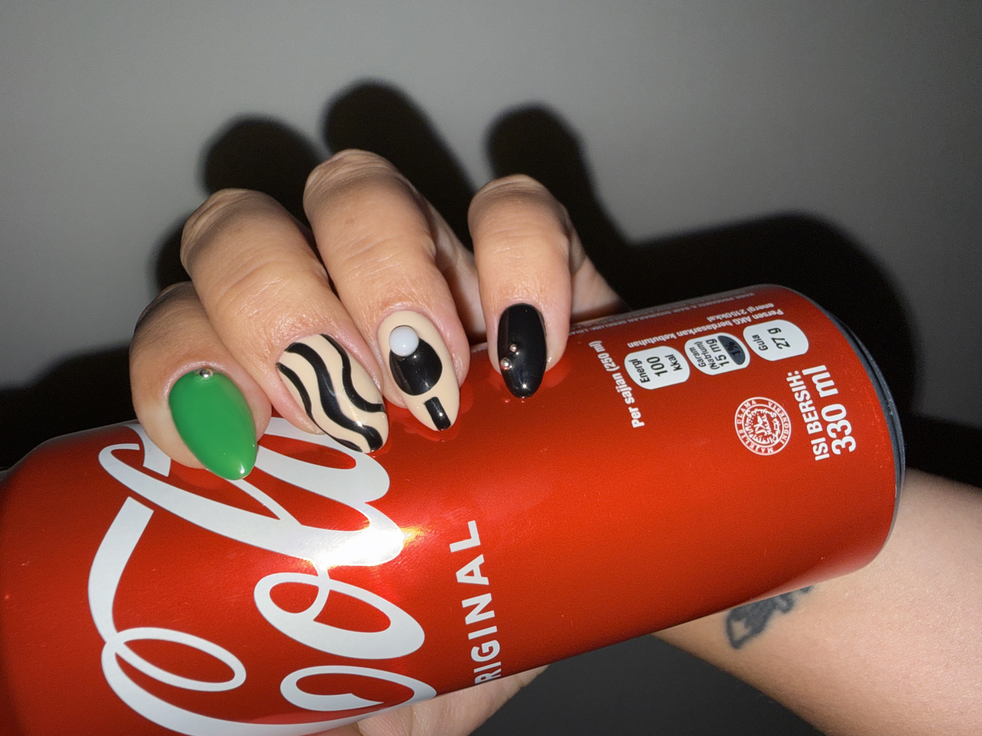 A close-up shot features a person's hand holding a red Coca-Cola can. The hand is adorned with unique nail art designs, including a green nail, a nail with black and beige zebra stripes, a beige nail with a black oval and white circle design, and a black nail with small silver accents. The Coca-Cola can displays the white logo, nutritional information in Indonesian, and the volume "330 ml." The text on the can also includes "ORIGINAL" and a Halal certification logo. The background appears to be a plain, muted wall, with a shadow visible above the hand. A tattoo can be seen on the forearm. Based on the Indonesian text on the can, the scene is likely in Indonesia. The can displays "Isi Bersih: 330 ml" which can be translated to "Net Volume: 330 ml". The nutritional information displays "Per sajian (250 ml)" which can be translated to "Per Serving (250 ml)", which is likely referring to 250 ml portions. Given the information on the can, it is highly unlikely the location is in North Kuta, Indonesia.