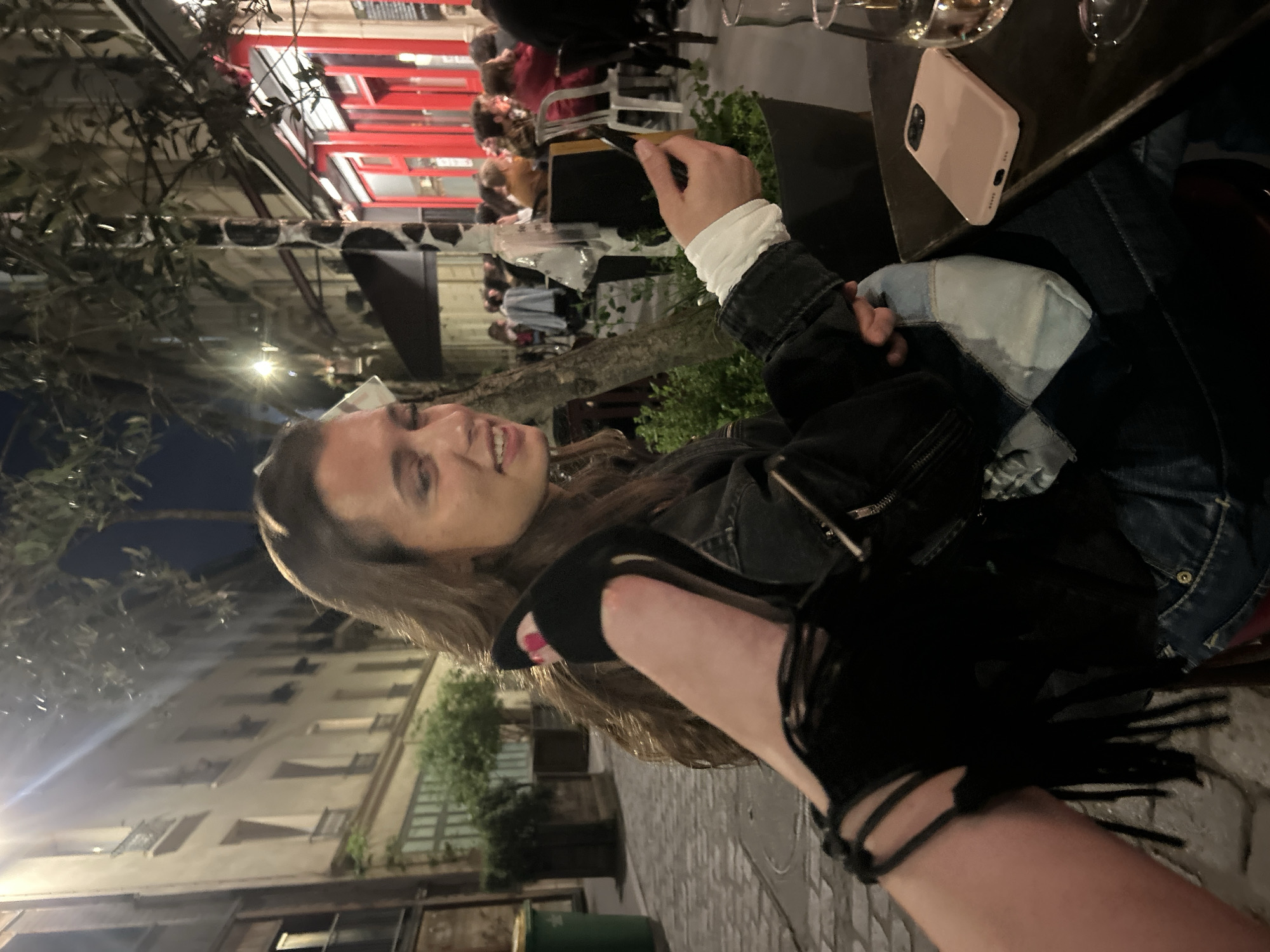A slightly tilted, full shot shows a fair-skinned woman, with light brown hair and a smile, holding a mobile phone outdoors in an urban environment. She is wearing a black jacket over a white shirt and a blue denim jacket. To the right of the woman is a brown table with a pink mobile phone and wine glasses on it. People are sitting at tables in the background, outside a building with a red awning. In the foreground, a person wearing a black sandal with fringe is holding their leg out, with the woman possibly taking a photo of the sandal. The pavement and buildings suggest the location is in a European city.