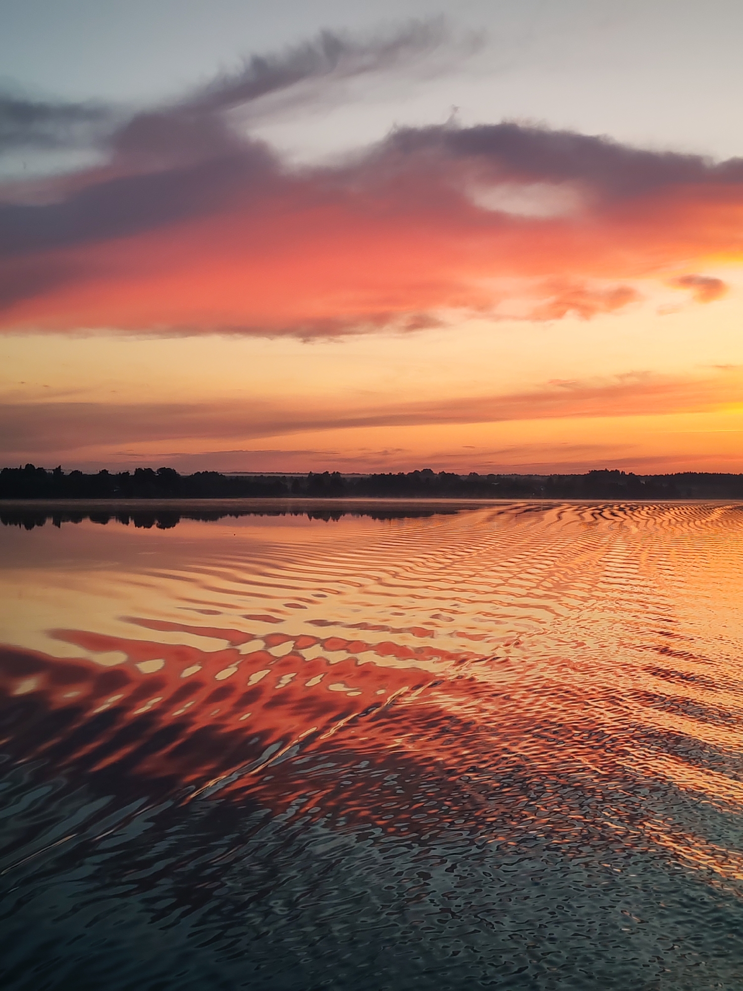 This image depicts a serene sunset over a body of water, likely a lake or a wide river, in or near the city of Плоски, Russia. The sky is ablaze with vibrant colors, transitioning from a pale grey at the top to soft oranges and fiery reds towards the horizon. Wispy clouds, tinged with purple and pink, stretch across the upper half of the frame. The water's surface is calm, reflecting the dramatic hues of the sky and creating a rippling pattern of light and shadow. In the distance, a dark silhouette of a treeline marks the edge of the land. The overall impression is one of tranquility and natural beauty, characteristic of a peaceful evening in a rural or semi-rural setting. There are no people or man-made objects prominently featured in the scene, emphasizing the natural landscape.