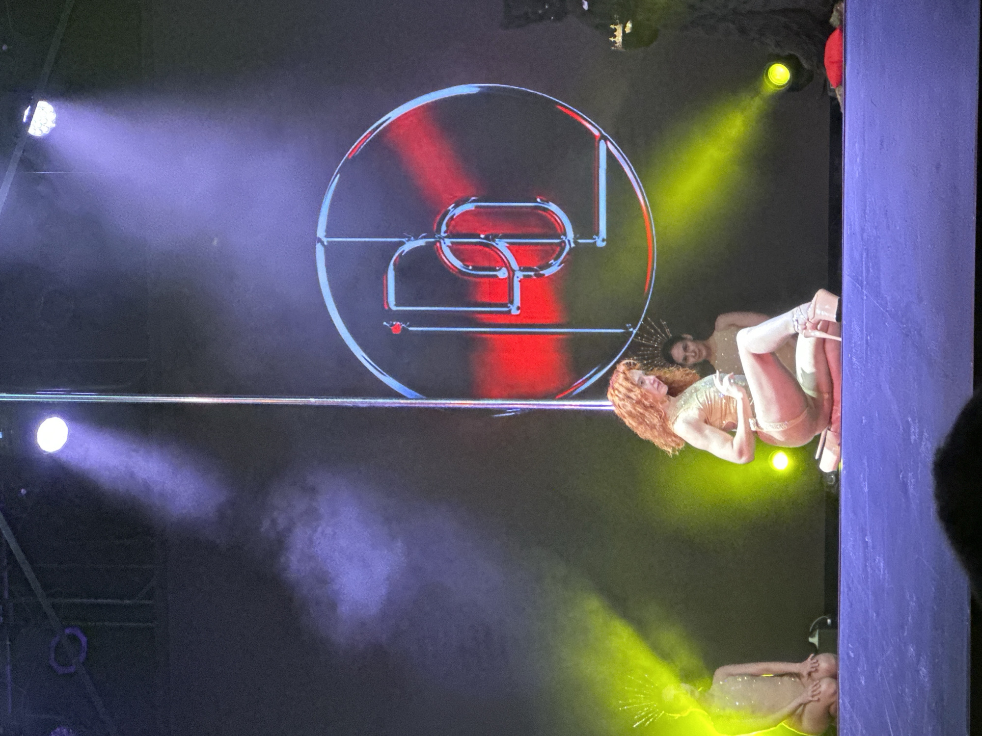 The image depicts a stage performance, possibly in Krasnoyarsk, Russia, given the presence of a Cyrillic "Д" incorporated into the circular logo at the back of the stage. Two female performers are visible under bright stage lights. One woman, with red-orange curly hair, is positioned near a vertical pole and is wearing a light-colored, possibly gold sequined, outfit and high heels. Another performer, appearing nude, is at the bottom of the stage. The backdrop behind them features a circular logo illuminated with red and blue lighting. The stage is dark with spotlights casting beams of light in blue and yellow. The edge of a structure, possibly part of the stage set, is seen on the right side of the image. The angle of the photograph is tilted, giving a skewed perspective of the scene.