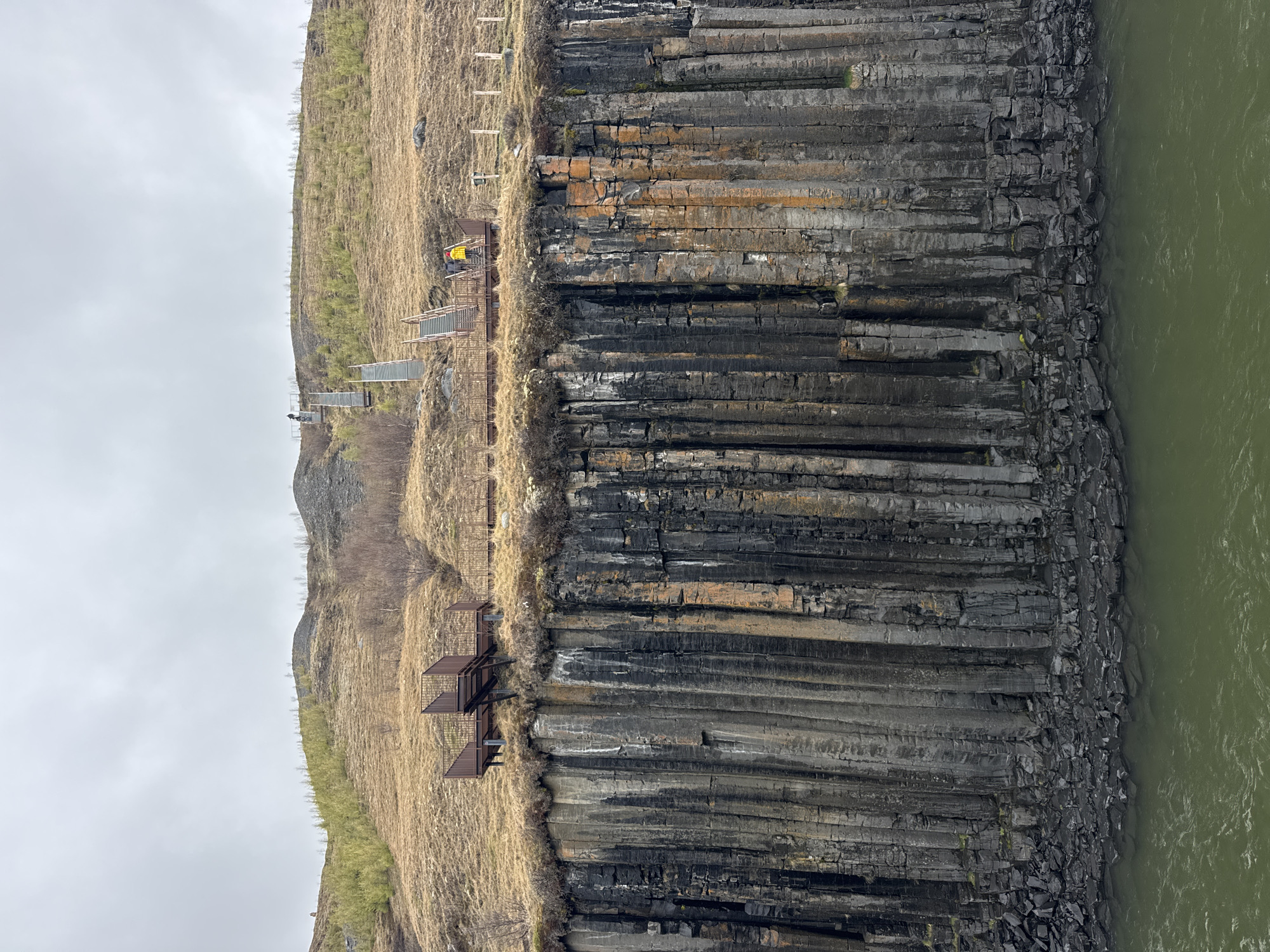 The image displays a dramatic natural landscape dominated by towering columnar basalt cliffs, seen from a perspective looking across a river. The cliffs are composed of dark grey and black hexagonal basalt columns, exhibiting various textures and shades, with some sections showing distinct orange, brown, and lighter grey weathering or mineral deposits. These impressive geological formations rise steeply from the banks of an olive-green river, which flows along the right side of the frame with subtle surface ripples.
Above the cliffs, a gently sloping hillside extends into the distance, covered in sparse, dry, golden-brown grass. A few thin, bare trees or shrubs are visible on the distant parts of the hill. Along the top edge of the cliffs, several man-made structures are visible, appearing to be viewing platforms and interconnected walkways. These structures are made of dark metal, suggesting robust construction for visitors.
On one of these platforms, a person wearing a bright yellow jacket or backpack is discernible, seemingly observing the vista. Another smaller figure can be seen on a more distant part of the walkway system. The overall scene suggests a destination for sightseeing or hiking, allowing visitors to experience the unique geological features. The sky is overcast and grey, indicating a cloudy daytime setting with diffuse light and no strong shadows. No visible text is present in the image.