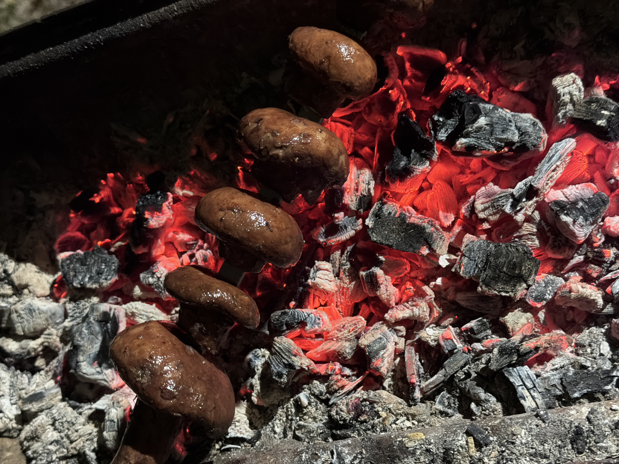 This is a close-up, overhead shot of several sausages cooking over glowing red embers and charcoal. The sausages are browned and appear to be glistening, indicating they are hot and likely juicy. They are skewered together, with some resting directly on the hot coals. The background consists of a dark, out-of-focus area, suggesting the photograph was taken at night or in low light conditions. The scene appears to be set outdoors, possibly in a backyard or campsite, given the presence of a grill or fire pit. There are no people visible in the image. The location is Unknown, Unknown.