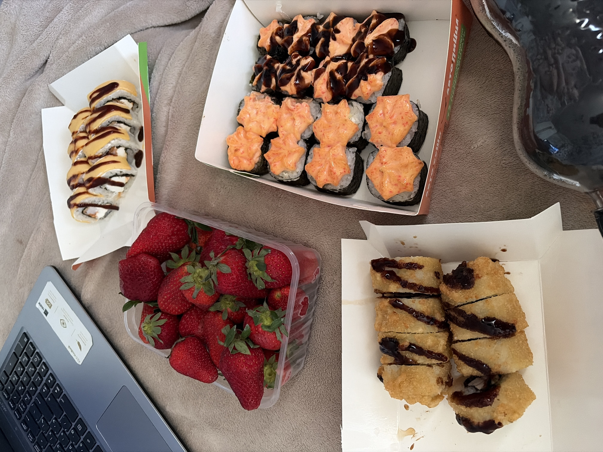 The image shows a laptop and several food containers arranged on a brown, textured surface, possibly a blanket or sofa. The food items appear to be sushi and strawberries. There are three boxes of sushi: one contains sushi rolls with a yellow topping, another with sushi topped with orange and dark brown sauces, and a third with fried sushi rolls drizzled with a dark sauce. A clear plastic container is filled with fresh strawberries. The laptop is open, revealing a keyboard and touchpad. Based on the container's text visible in a cropped image, the food might have been purchased from "Auto Pizza", a brand with known outlets in Новинки, Russia.