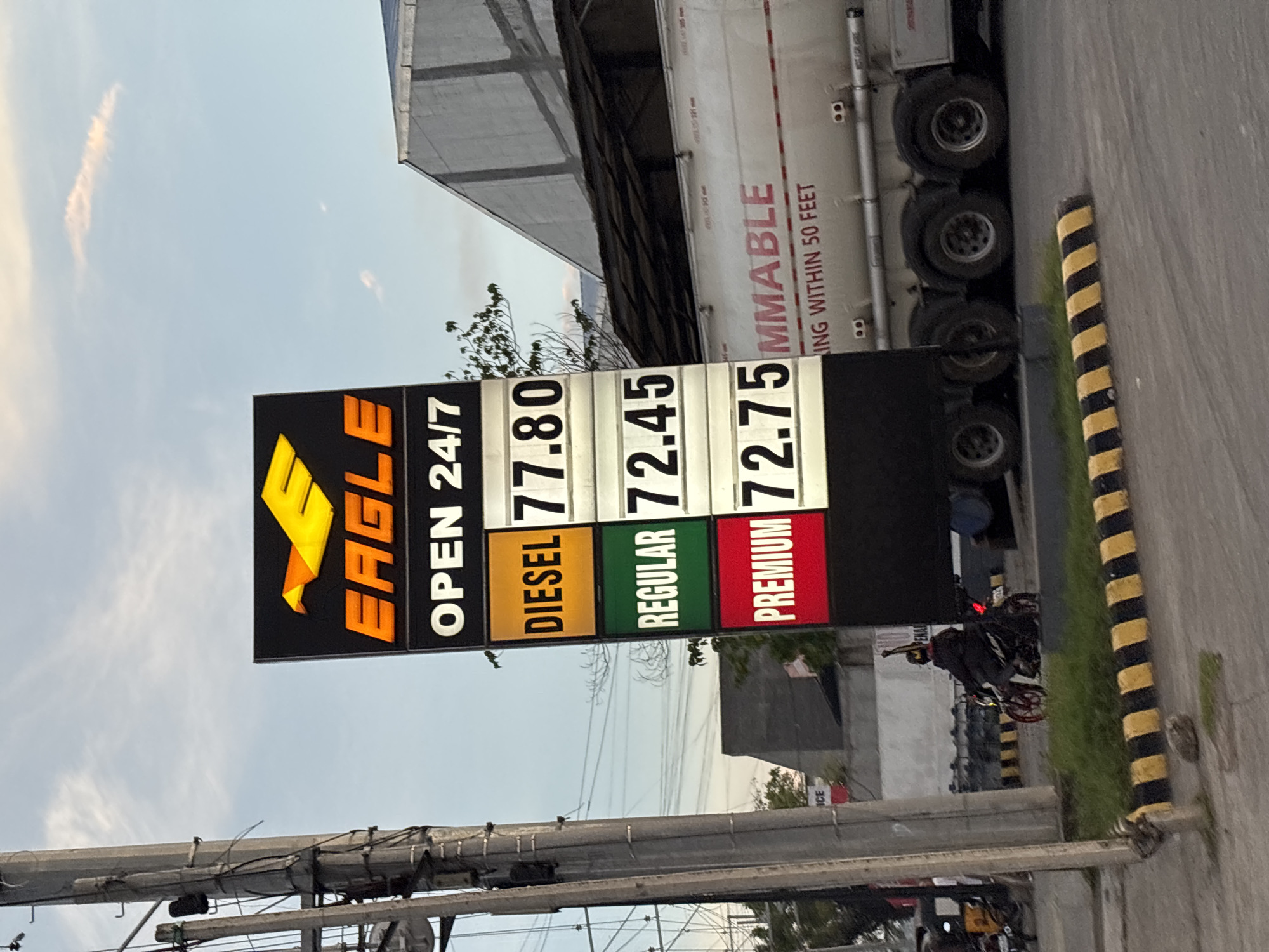 The image captures a gas station sign in Davao City, Philippines, on a clear day with a pale sky and a few wispy clouds. The sign displays fuel prices for "DIESEL" at 77.80, "REGULAR" at 72.45, and "PREMIUM" at 72.75. Above these prices, it reads "EAGLE OPEN 24/7". To the right of the sign, a large white tanker truck with the word "FLAMMABLE" printed in red is visible, along with text indicating to keep a distance of "WITHIN 50 FEET". In the foreground, a person on a red and black motorcycle is stopped at the intersection, facing away from the camera, with their left arm raised. A utility pole with electrical wires stands prominently on the left side of the image. Yellow and black striped barriers are present on the road.