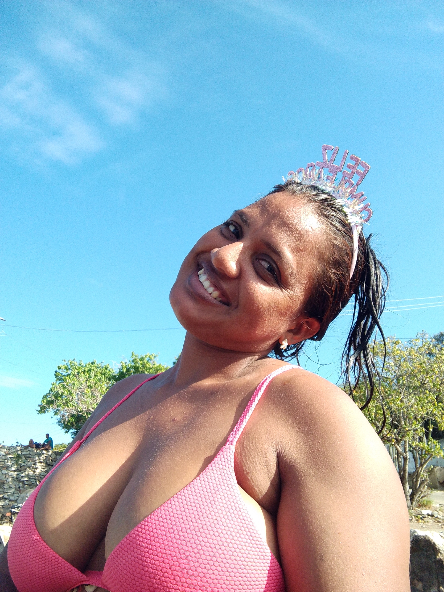 An outdoor, low-angle, close-up, selfie-style photograph shows a dark-skinned woman wearing a pink bikini top and a sparkly tiara with the words "THE BEST" on it. Her head is tilted to her left, and she is smiling with her mouth open, revealing her teeth. Her hair is wet and pulled back into a ponytail. She is wearing a small white flower earring in her visible ear. The background consists of a bright blue sky with a few wispy white clouds, and some greenery and a stone wall behind her. The overall impression is that of a woman enjoying a sunny day, possibly celebrating a birthday. The location is unknown.