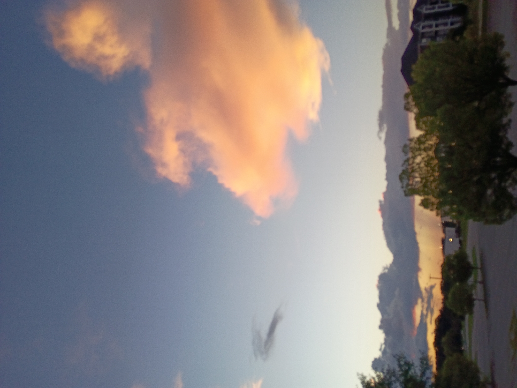 The image depicts a cloudy sky at sunset over a suburban area. The sky is filled with large, dramatic clouds, some of which are illuminated with hues of orange and pink from the setting sun. Below the sky, silhouetted trees and buildings can be seen. The foreground shows a paved area, possibly a parking lot, with some trees scattered around. The location is identified as Nicholasville, United States of America.