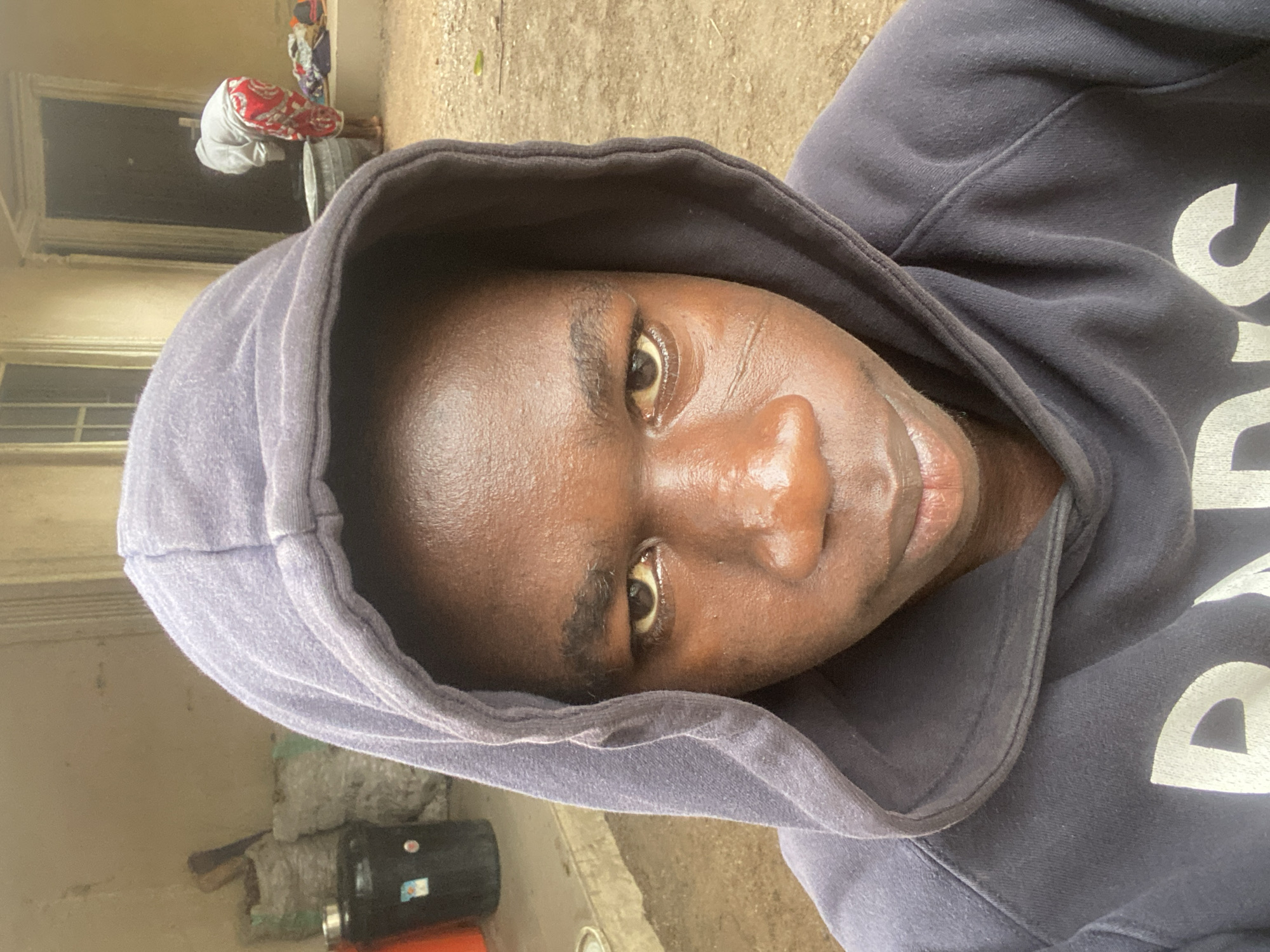 A forensic analysis of the image reveals an outdoor scene in Mararaba, Nigeria. In the foreground, a young individual with dark skin, wearing a dark hooded sweatshirt with partial white lettering ("D" and "S") visible on the chest, faces directly towards the camera. The individual exhibits a serious expression, with dark eyes that appear moist or shiny. Notable facial scars are present on the individual's left cheek (viewer's right).
The background depicts a residential or utilitarian outdoor area. To the left of the foreground subject, a light-colored wall stands adjacent to several white sacks, possibly filled with materials. A black drum with a metallic lid and a sticker is positioned in front of the sacks, with an orange object partially visible nearby. The ground in this section appears to be a concrete or paved surface.
To the right, a second individual is visible in the mid-ground, bent over and facing away from the camera. This person wears a light-colored top and red and white patterned pants, and appears to be interacting with a large, grey, cylindrical basin on the unpaved, dirt or gravel ground. Additional items are scattered near this person. A light-colored building with a dark doorway and a window frame forms the backdrop on this side. The overall lighting indicates natural daylight, consistent with an outdoor setting.