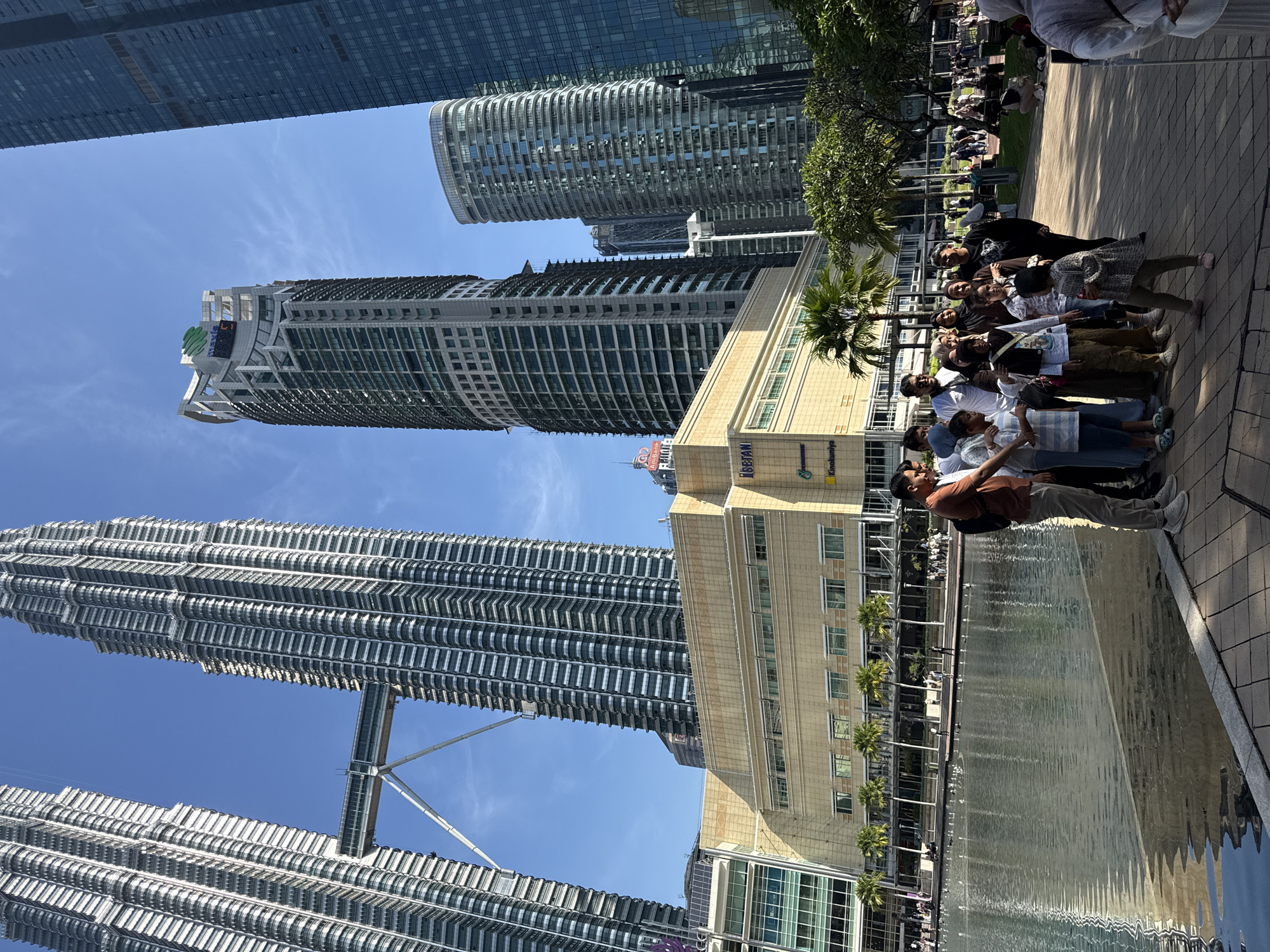 The image shows a group of people posing for a photo in Kuala Lumpur, Malaysia. The iconic Petronas Twin Towers are prominently featured in the background. Adjacent to the towers is a building with visible signage including 'Koolunya' and 'PETRONAS'. Other high-rise buildings are also visible. The group of people is lined up on a paved area next to a body of water. Some people are sitting on a grassy area further back. The sky is clear and blue. The photo is taken on a sunny day.