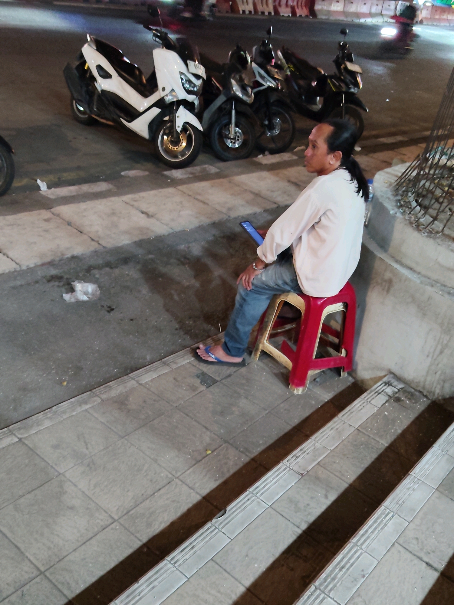 A man with his hair tied in a ponytail sits on a red plastic stool, holding a smartphone. He is wearing a white long-sleeved shirt and blue jeans. He is wearing flip-flops. The man is sitting on a sidewalk next to a concrete wall. In the background, there are several motorcycles parked on the street. The scene appears to be in an urban area at night, likely in Purwakarta, Indonesia. The lighting suggests it is nighttime, with streetlights or other artificial illumination. There is some debris on the ground, including a crumpled plastic bag. The ground is paved with tiles.