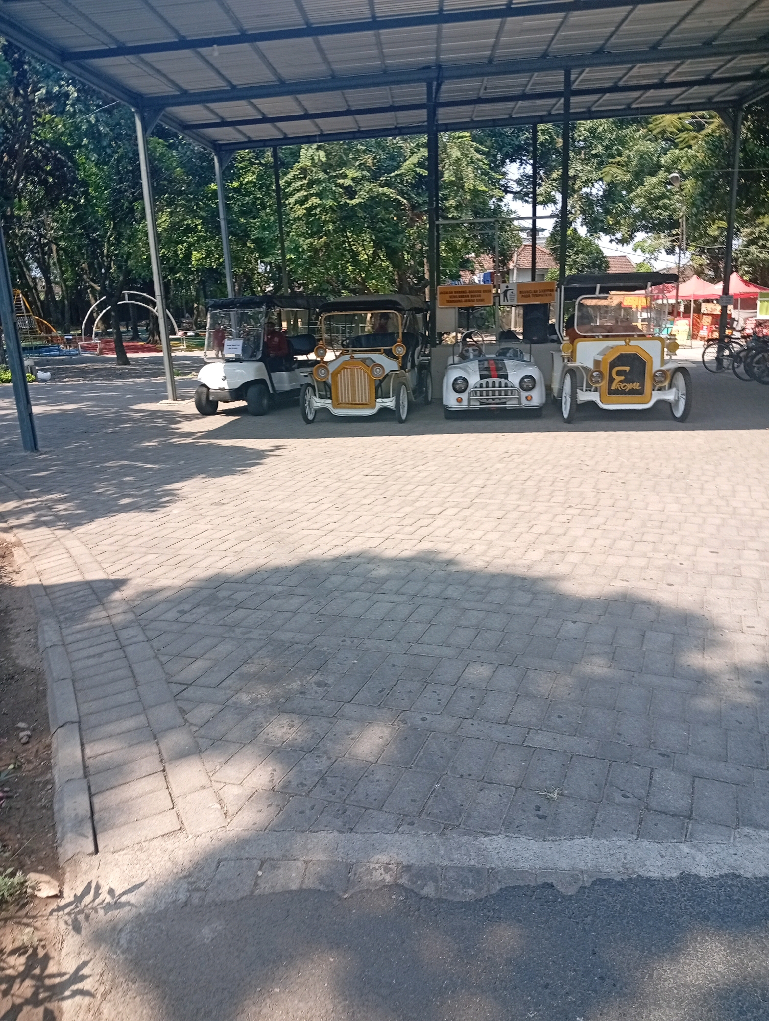 The image shows a shaded parking area with four small, vintage-style cars parked under a metal canopy. The cars are of varying colors, including white, yellow, and red. Two of the cars are open-top, while the other two have canopies. The ground is paved with interlocking bricks. In the background, there are trees, a playground, and some buildings with red roofs. There are also bicycles parked to the right of the cars. The overall scene suggests a recreational area or a tourist attraction. The location is identified as Pandaan, Indonesia.