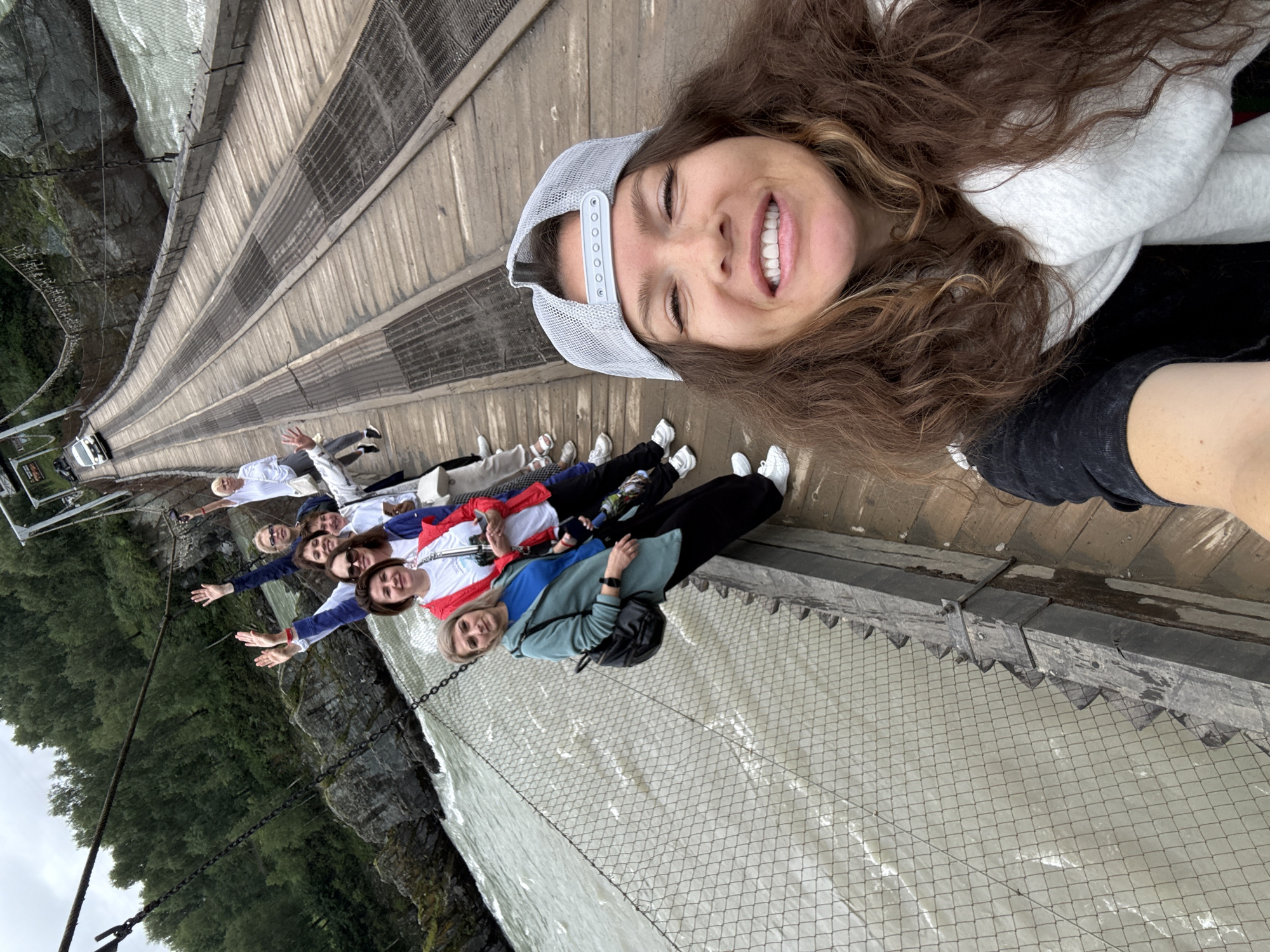 A group of eight people are standing on a wooden suspension bridge over a turbulent river. The bridge has a metal mesh walkway and a wire fence along the sides. The people are arranged in a line, with the person taking the selfie at the front. The person in the foreground is a woman wearing a grey baseball cap, a white scarf, and a black t-shirt. She is smiling and looking at the camera. The other seven people are behind her, and most of them are waving. They are dressed in casual clothing. The river is a murky grey-green color, and the surrounding landscape is characterized by rocky cliffs and dense green forest. A white car is visible on the bridge in the background. The sky is overcast. The scene appears to be in a natural, outdoor environment. The location is specified as Elekmonar, Russia.