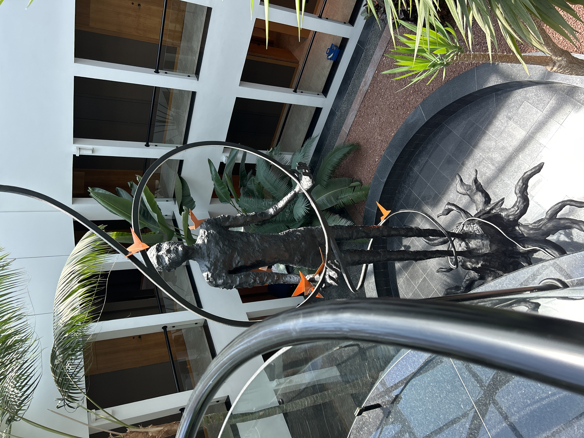 A full shot from a high angle captures a modern interior courtyard with a dark metal sculpture of a person standing in the center. The sculpture is surrounded by lush green tropical plants, including palm trees and ferns. The courtyard has a circular black tiled floor with a dark red gravel border. Surrounding the courtyard are multiple levels of balconies with wooden doors and black metal railings. A curved black metal railing dominates the foreground, obscuring some of the scene. The walls of the building are white, and the lighting suggests a bright, airy space.