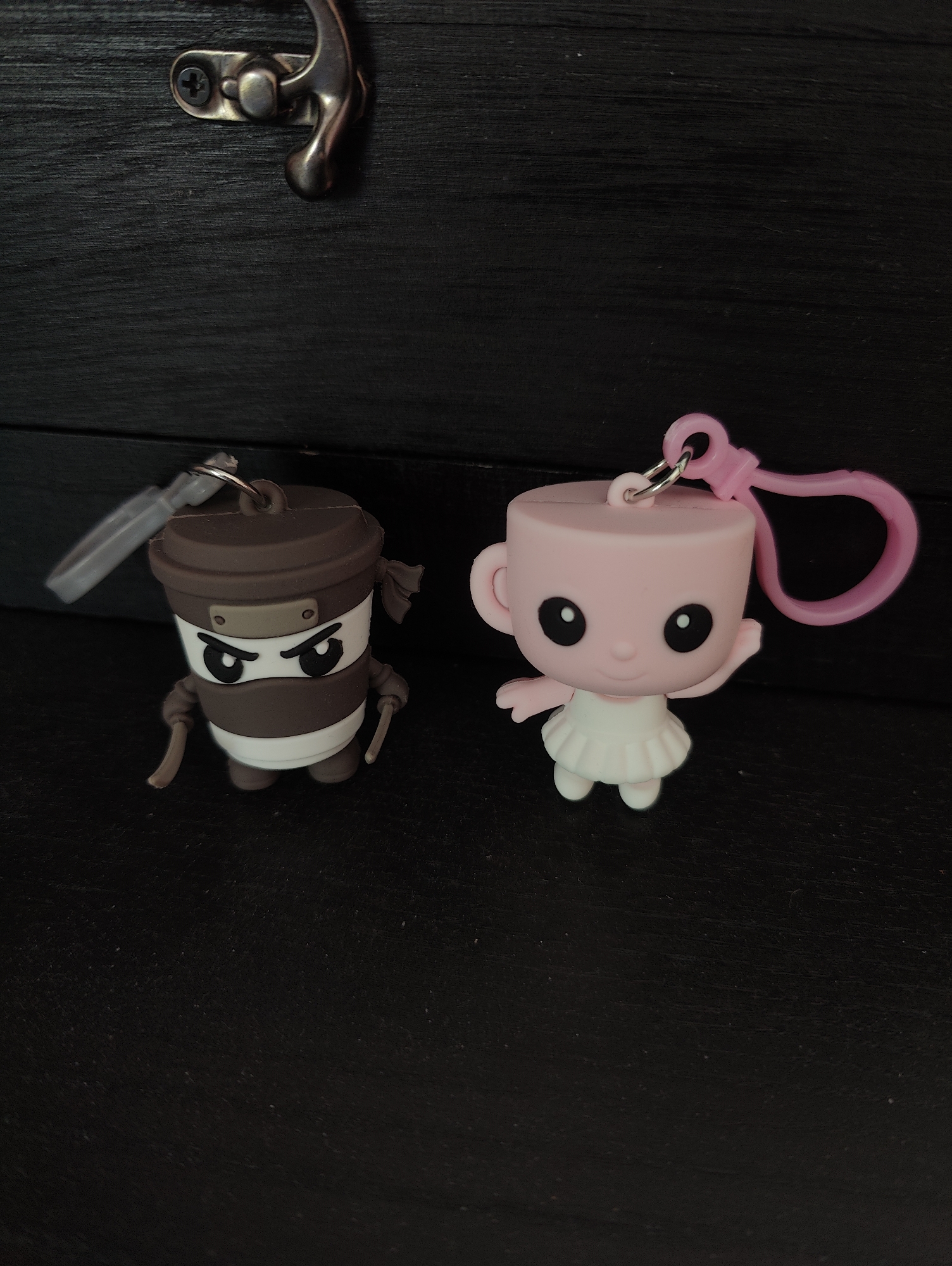 The image shows two cartoon character keychains resting on a black surface. The keychain on the left is brown and white, resembling a coffee cup ninja with a headband and katanas. The keychain on the right is pink, resembling a teacup ballerina with a tutu and a pink clasp. The surface appears to be a dark, possibly wooden, texture. In the upper left corner, there's a partial view of a metal hinge or clasp, attached to what seems to be a dark wooden or similarly textured surface. The location of the city of Ipswich, United Kingdom, is not discernible from the image.