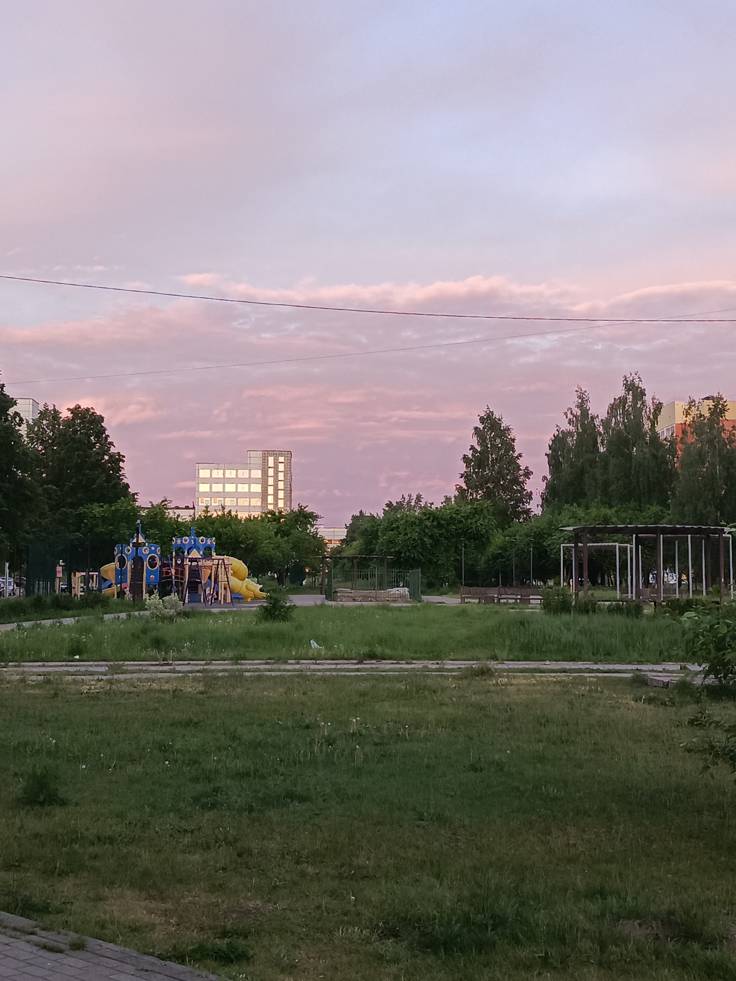 The image depicts an urban scene in Chaikovsky, Russia. The foreground is dominated by a grassy field, slightly overgrown with patches of weeds. A low concrete barrier separates the field from the viewer. In the middle ground, there's a playground with colorful blue and yellow play structures. A building with multiple windows reflecting the evening light is visible in the background, nestled among trees. There's also a covered seating area or pergola to the right. Above, the sky is a gradient of pink and purple, suggesting either dawn or dusk. A power line stretches across the upper portion of the image. No people are clearly visible.