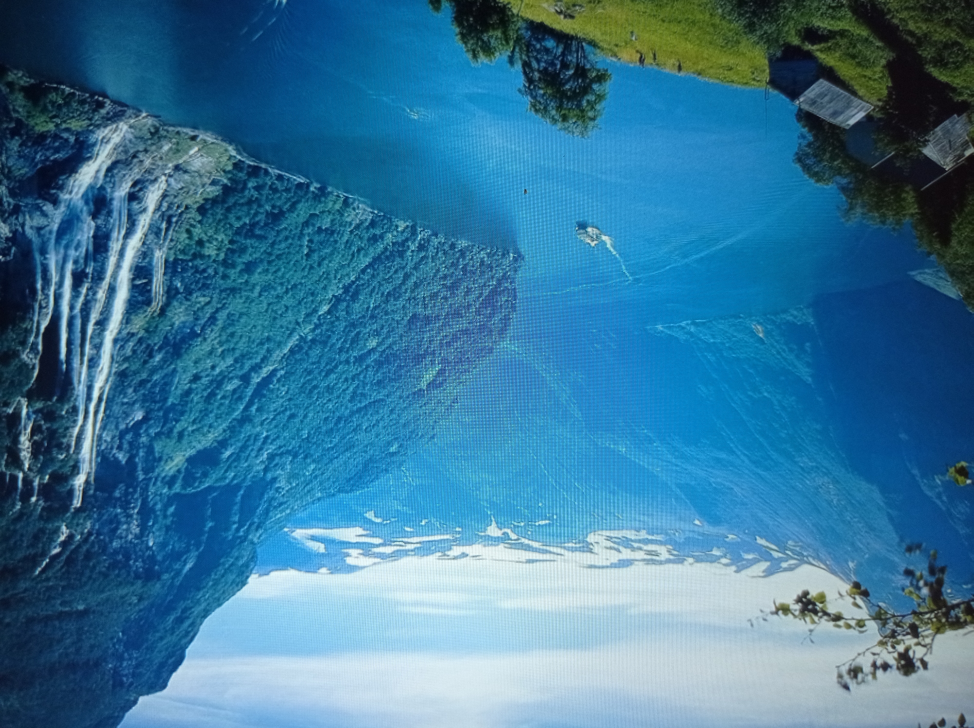 The image displays a majestic fjord with steep, lush green mountains on either side. A deep blue body of water, likely a lake or sea, fills the center of the frame, with a small boat or ship navigating its surface. Numerous waterfalls cascade down the rocky slopes of the mountain on the right, adding to the dramatic scenery. The upper reaches of the mountains are capped with snow. In the foreground, on the left side of the image, several rustic wooden cabins are nestled on a grassy hillside. A few individuals can be seen walking on the hillside. The sky above is a clear, bright blue with some scattered clouds. The scene appears to be a natural landscape, likely in a mountainous region known for fjords and waterfalls. This image does not depict any elements or scenery associated with the city of Mukachevo, Ukraine.